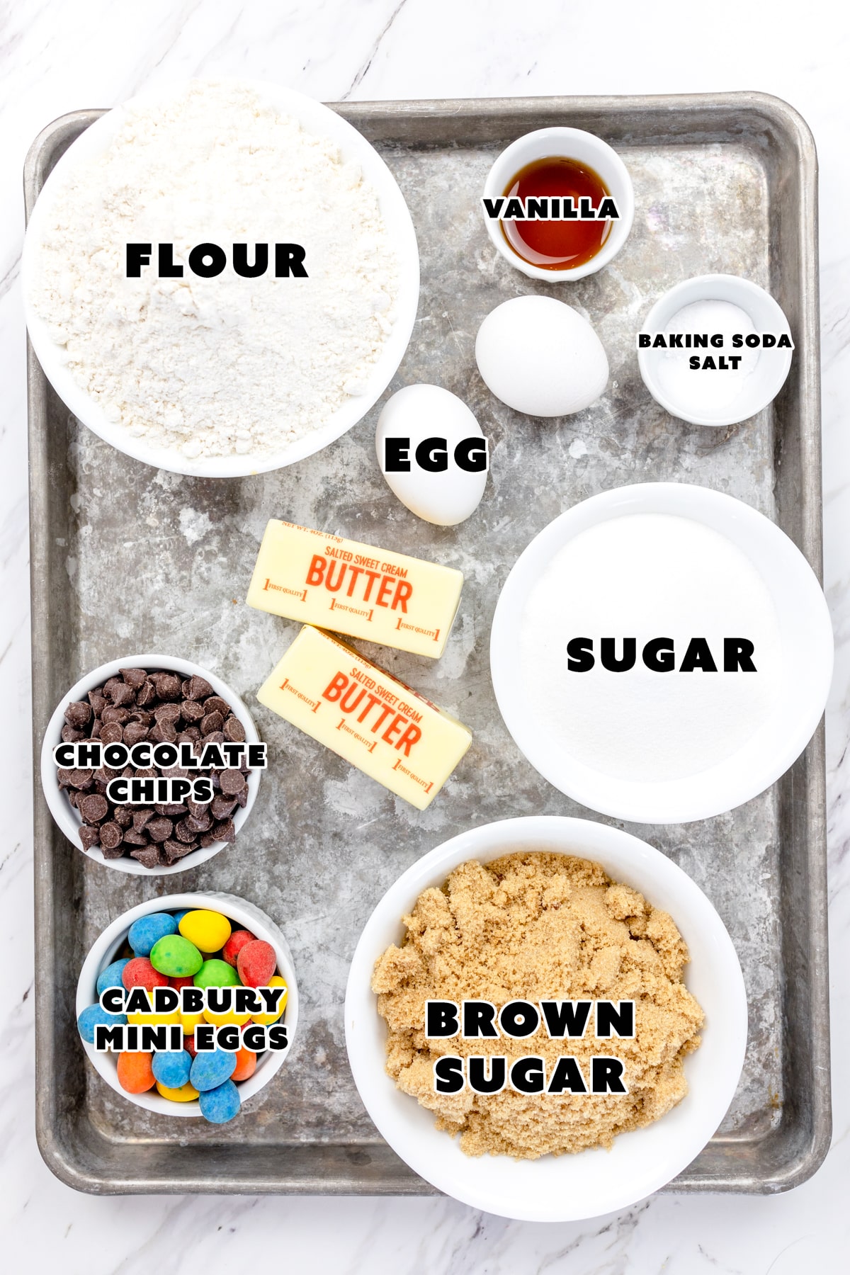 Top view of ingredients needed to make Cadbury Mini Egg Cookies in small bowls on a baking tray with labels on them. 