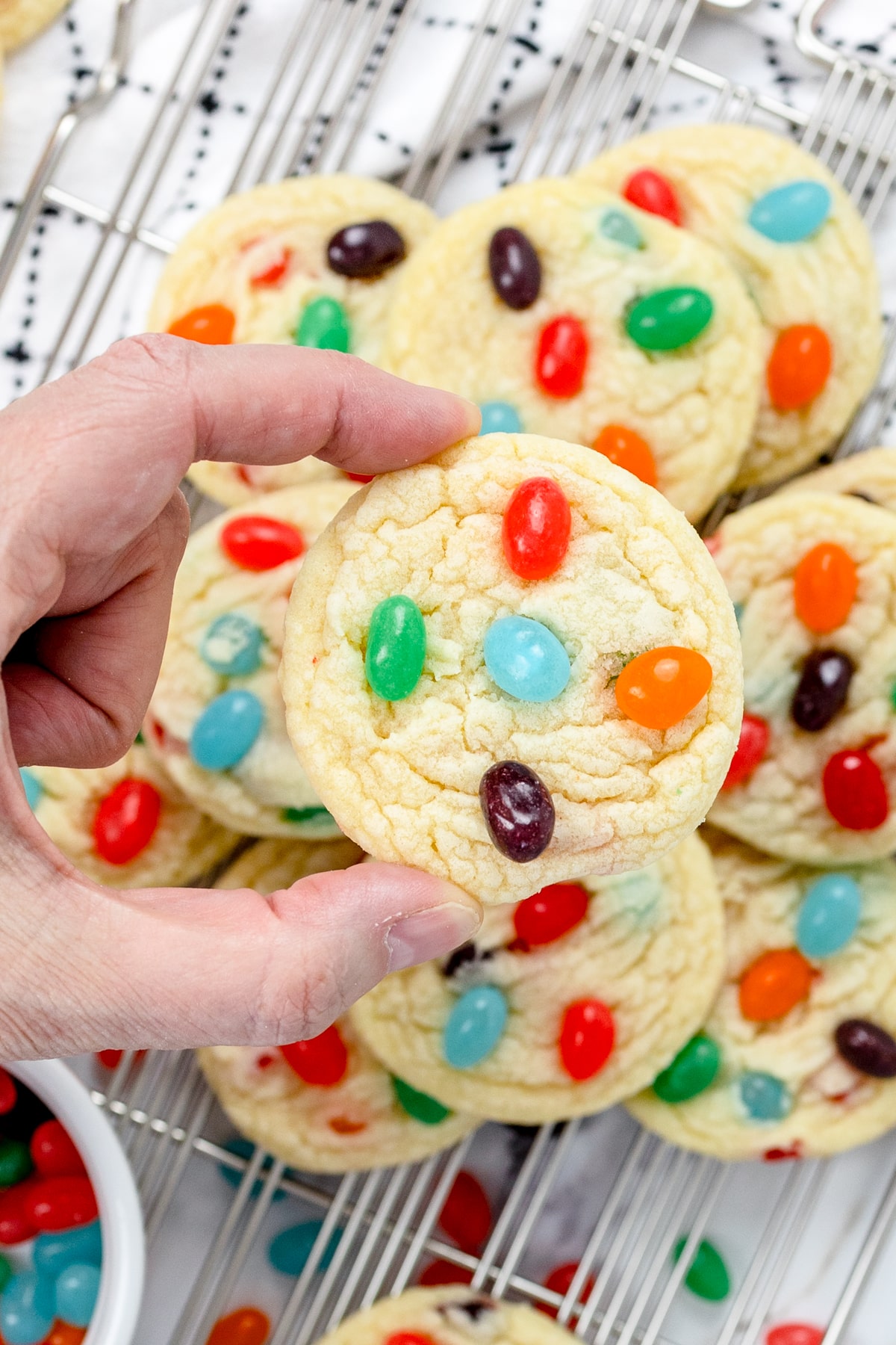 Top view of someone holding a Jelly Bean cookie in mid-air above a rack filled with cookies. 