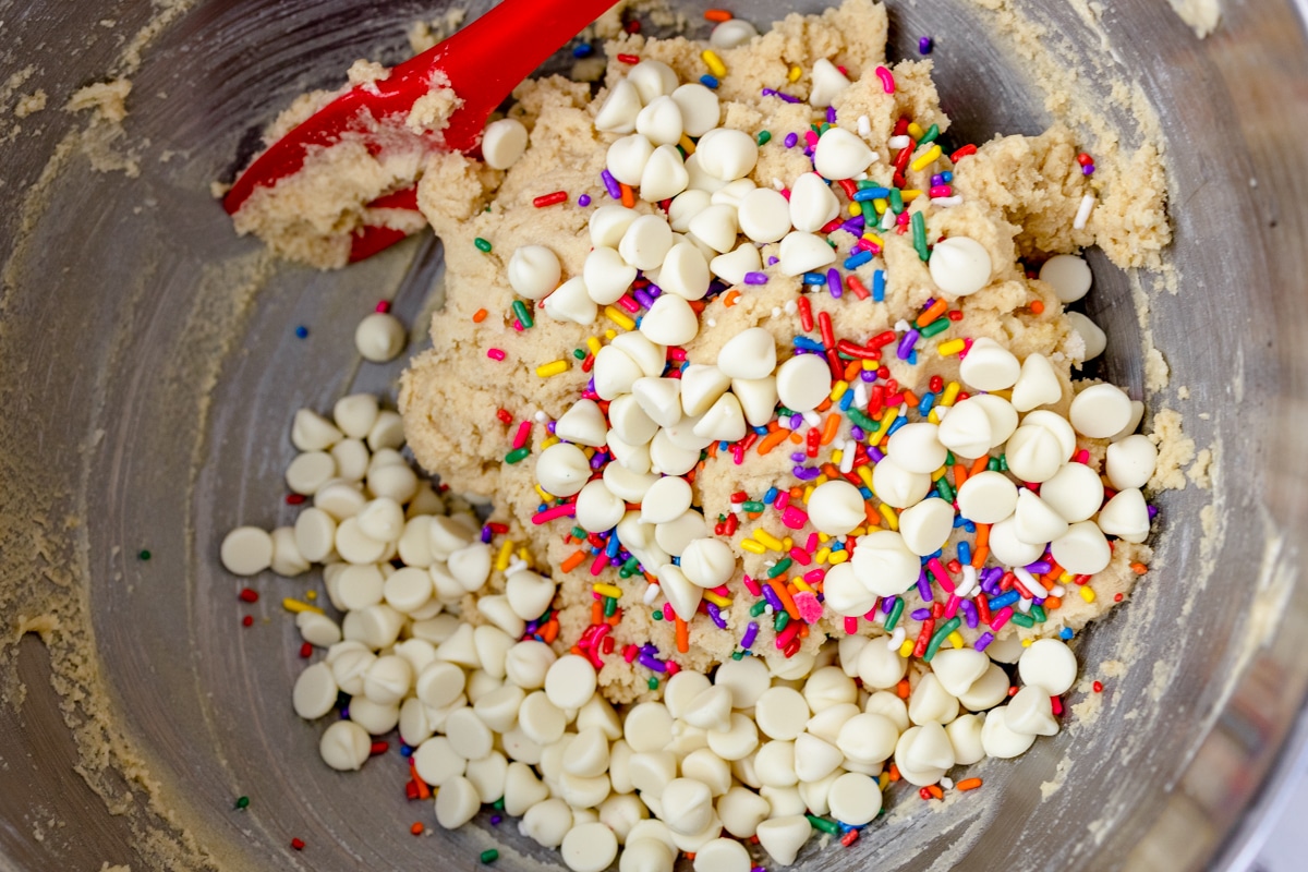 Top view of cookie dough in a mixing bowl with rainbow sprinkles and white chocolate chips on top. 