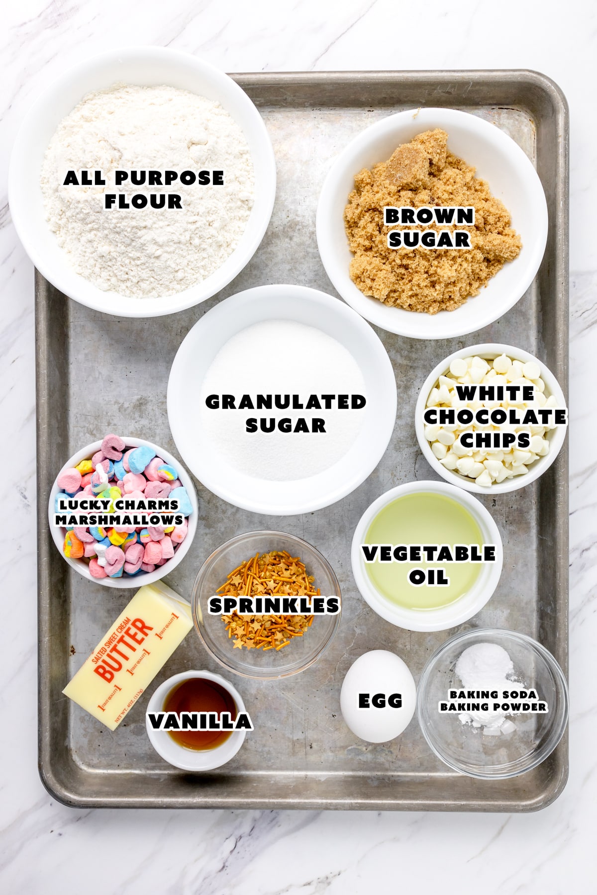 Top view of ingredients needed to make Lucky Charms Cookies in small bowls on a baking tray.