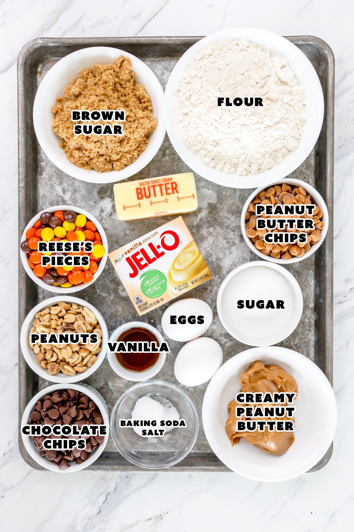 Top view of ingredients needed to make Reeses Pieces Cookies in small bowls on a baking tray with labels on them.
