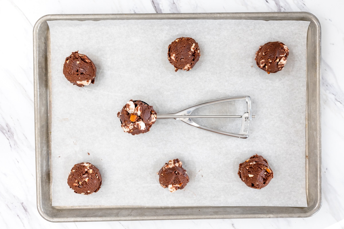 Top view of rocky road cookie dough balls on a baking tray with a cookie scoop in the middle of the tray. 