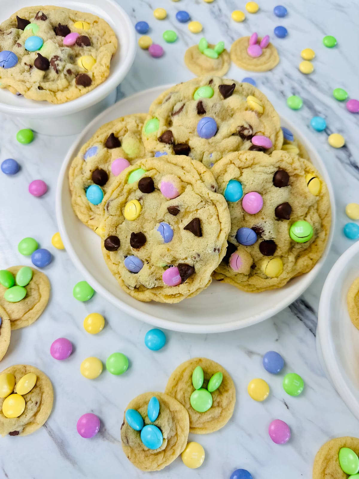 Top view of a plate filled with Easter M&M cookies, with pastel M&Ms on the table around it, and also some mini bunny cookies next to it. 