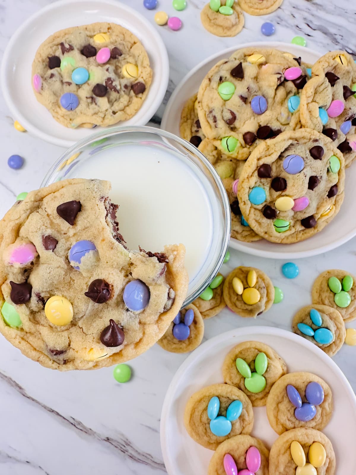Top view of an Easter M&M Cookie sitting on the rim of a glass of milk, with a plate of cookies next to it. 