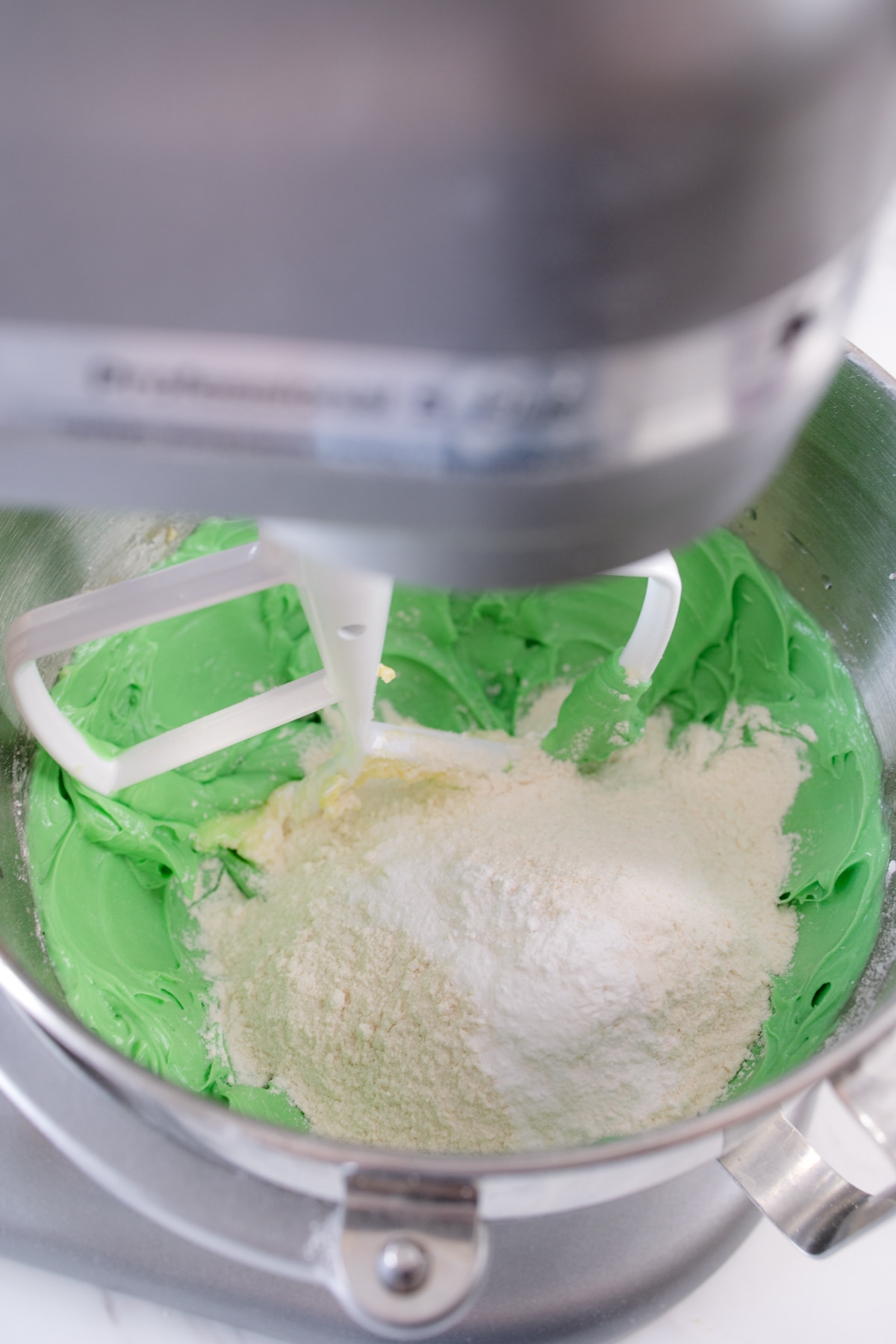 Close up of stand mixer with a green cookie dough mixture in it with flour being added to it. 