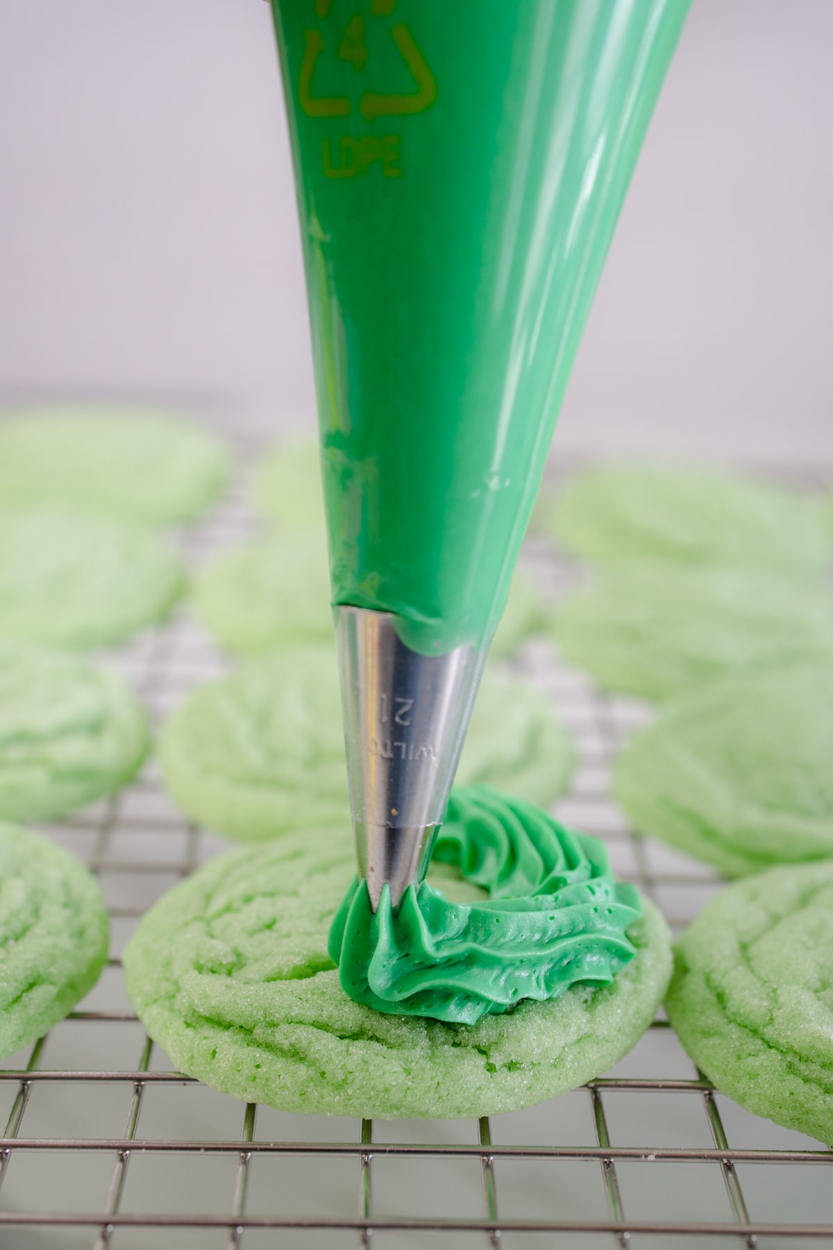 Close-up of a piping bag piping green frosting onto a green cookie sitting on a wire rack. 