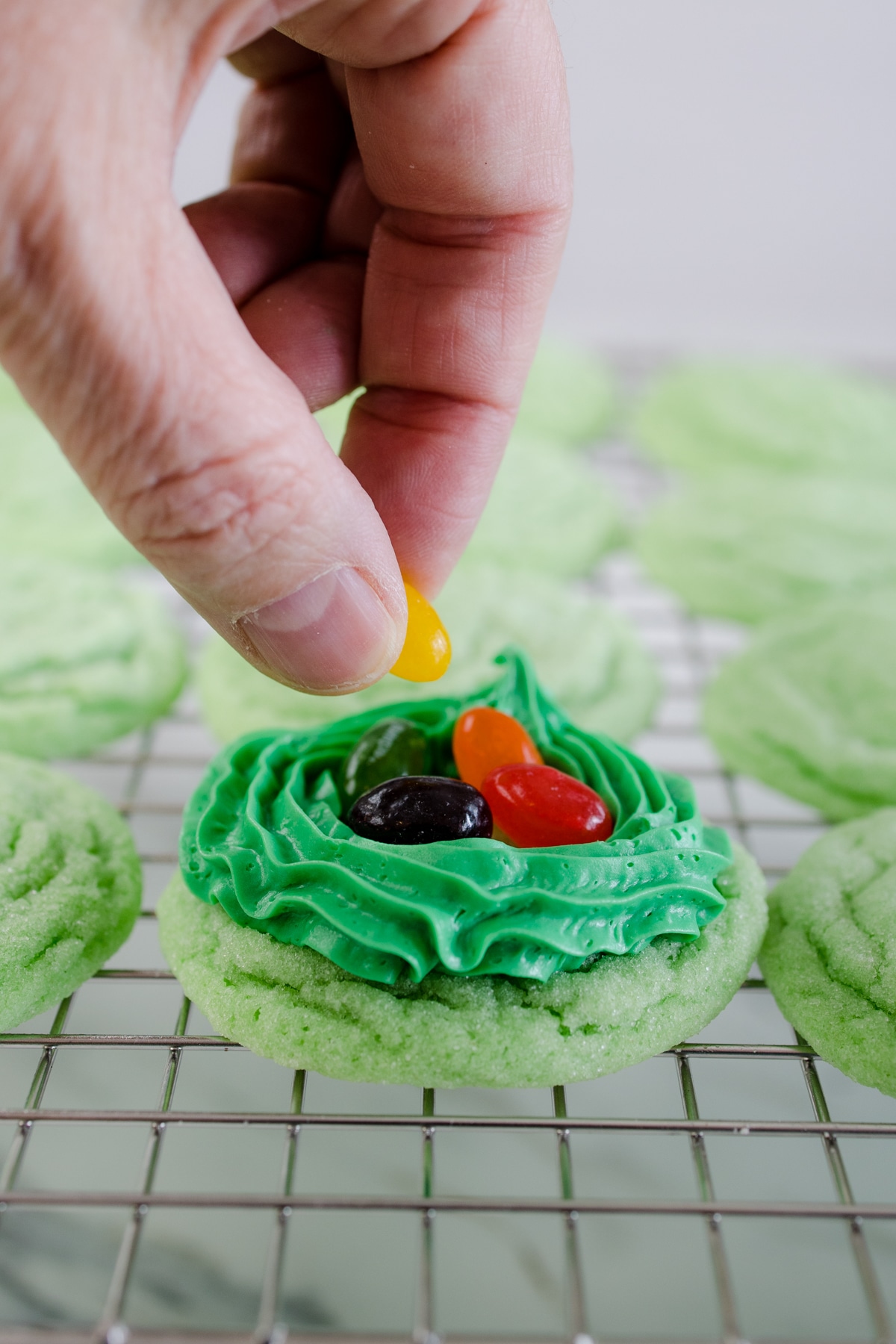 Close up of a hand putting jelly beans into the green frosting 'nest' on top of a green cookie on a wire rack. 
