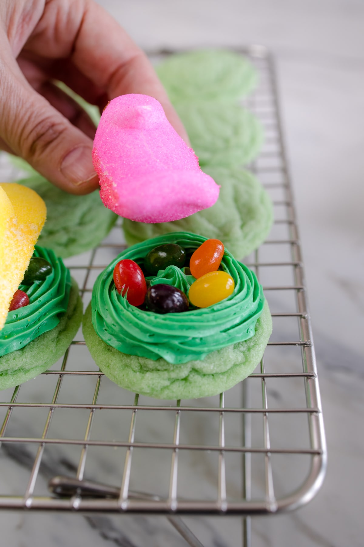 Close up of a pink chick Peep marshmallow being put on top of jelly beans on a green frosting 'nest' on top of a green cookie on a wire rack.