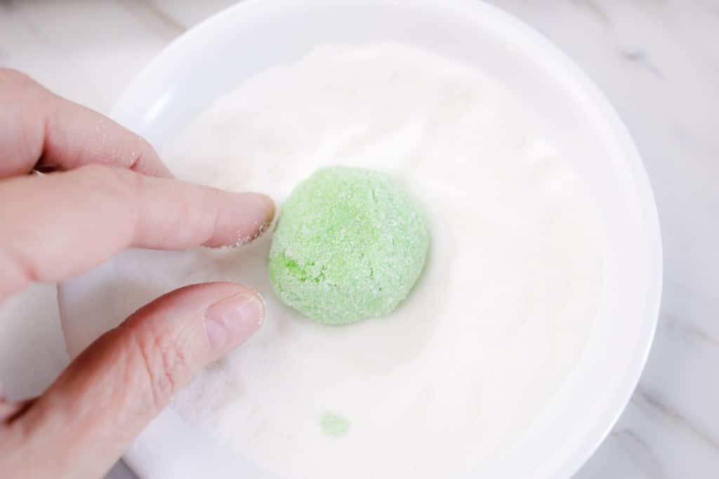 Close up of fingers rolling a small green ball of cookie dough into a bowl of granulated sugar to coat it.