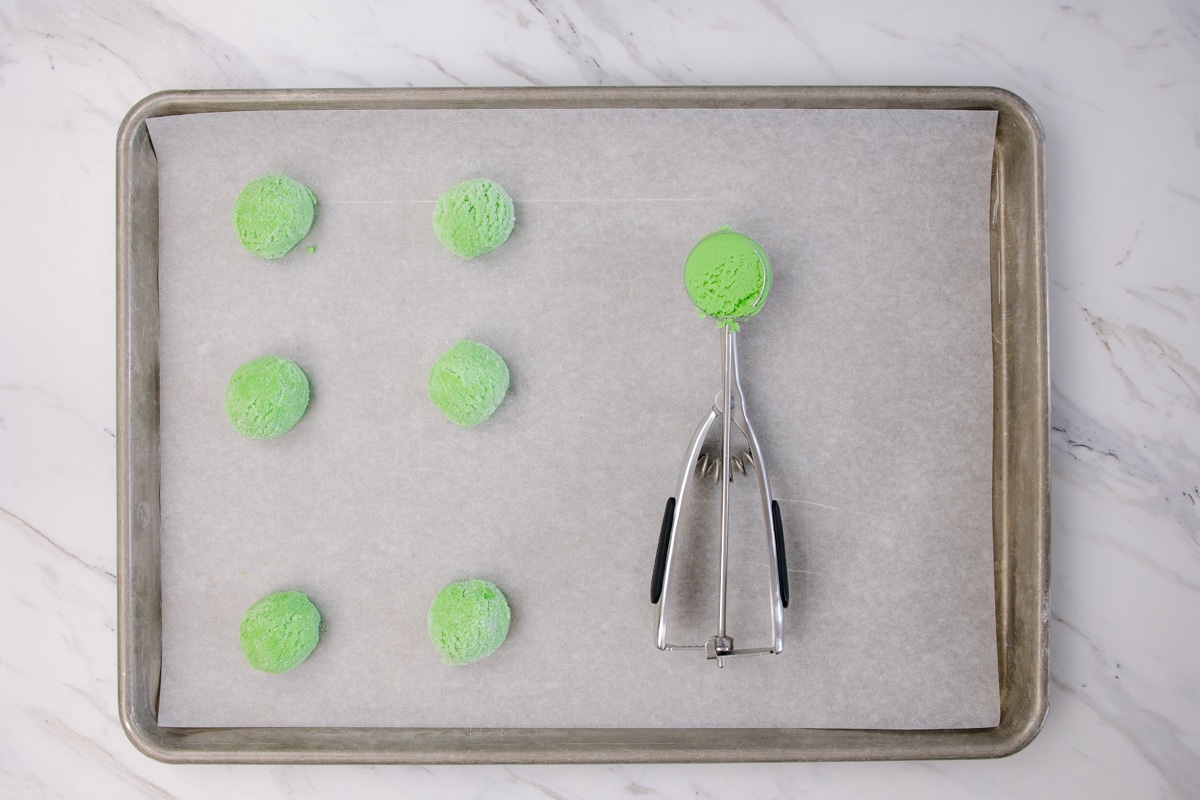 Top view of baking tray with a small cookie scoop lying on it, with a small ball of green cookie dough in it, next to six small green balls of dough. 