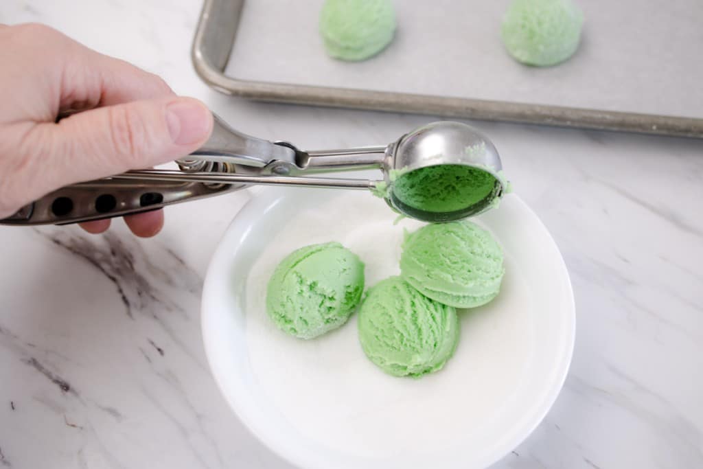 Close up of a small cookie scoop putting three small green balls of cookie dough into a small bowl of granulated sugar. 