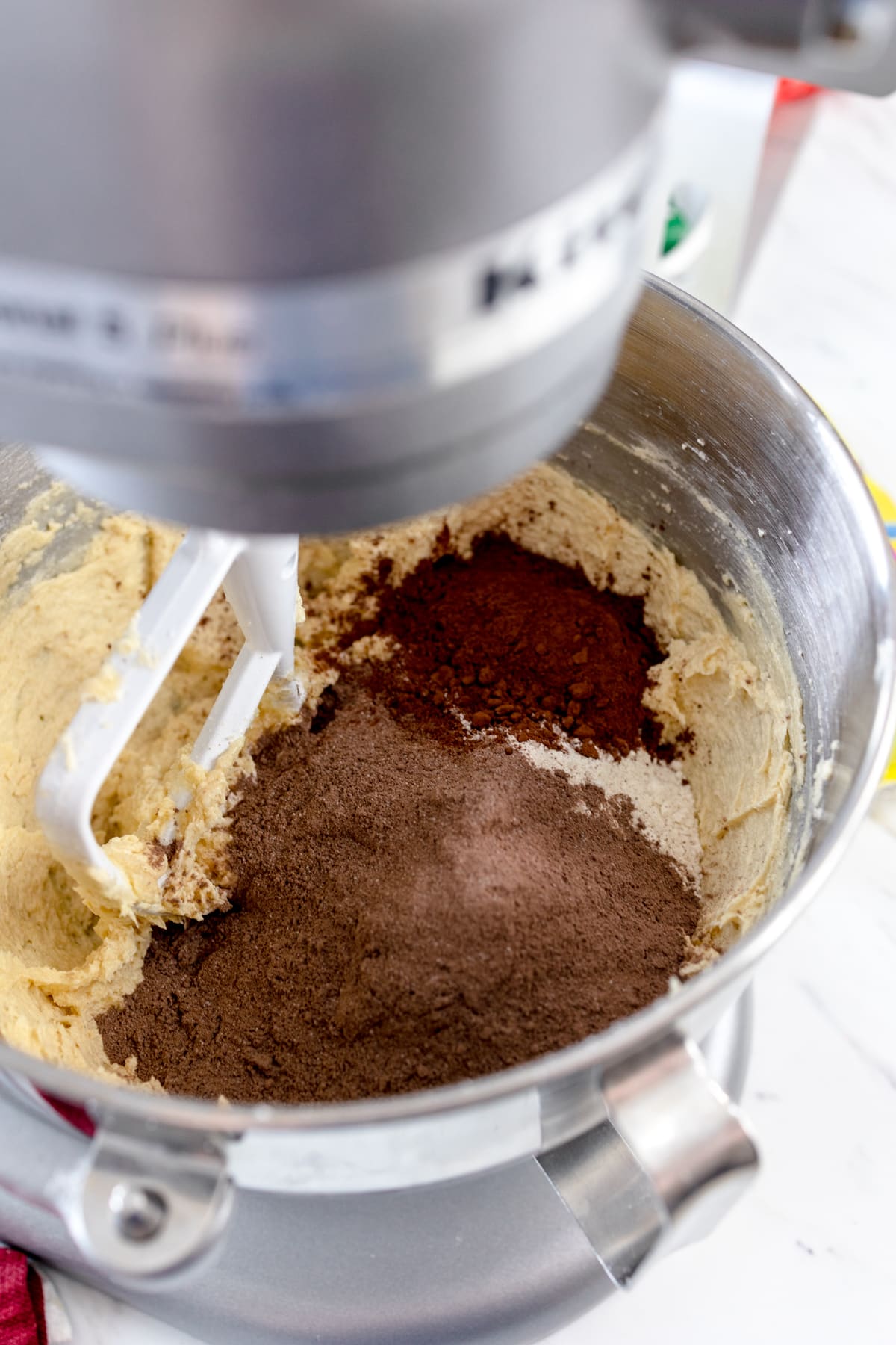 Close up of the bowl of a stand mixer with cookie dough mix in it and cocoa powder being added on top.