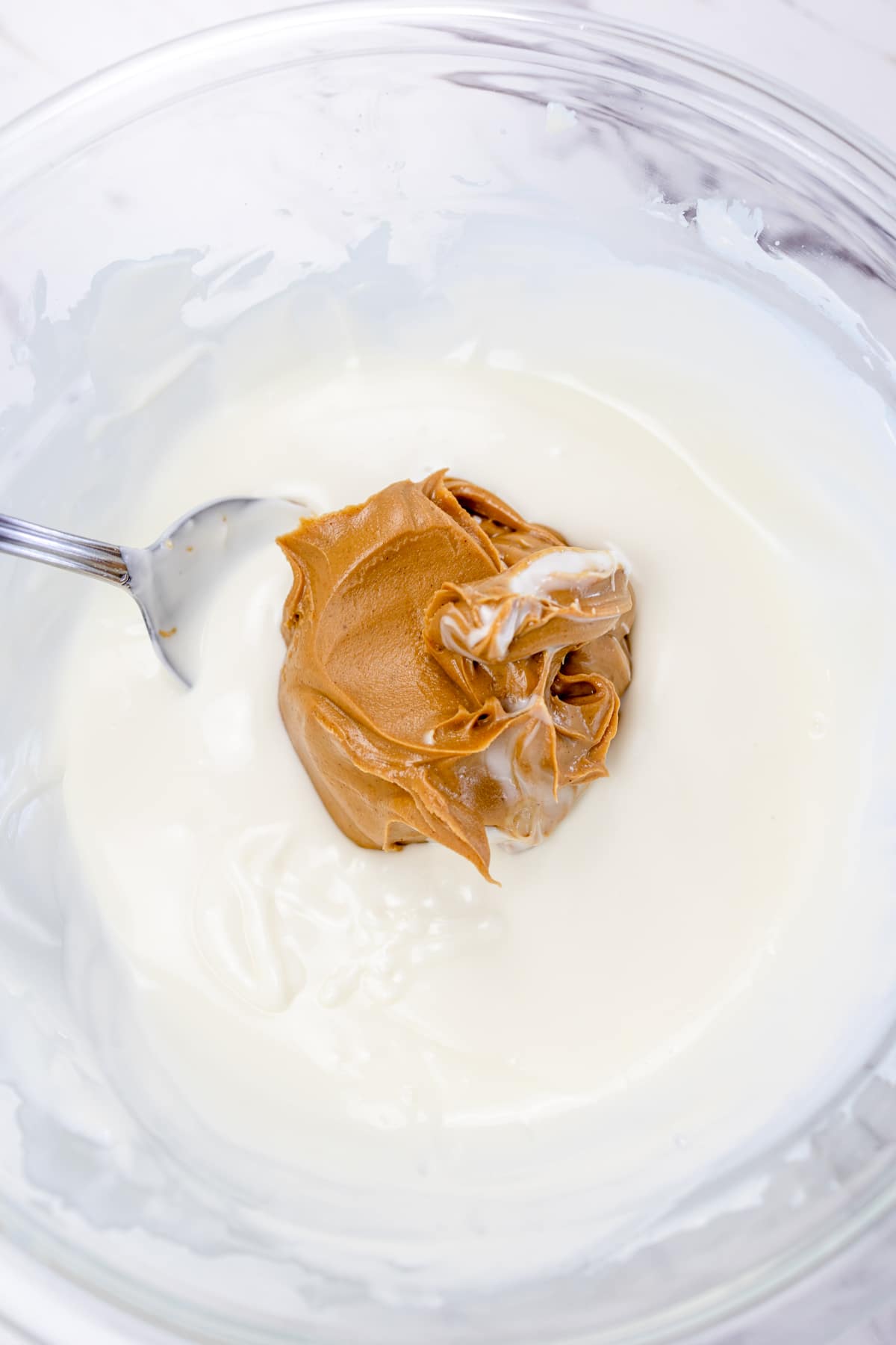 Top view of a glass mixing bowl with a melted white mixture in it and a dollop of peanut butter being added in with a spoon.