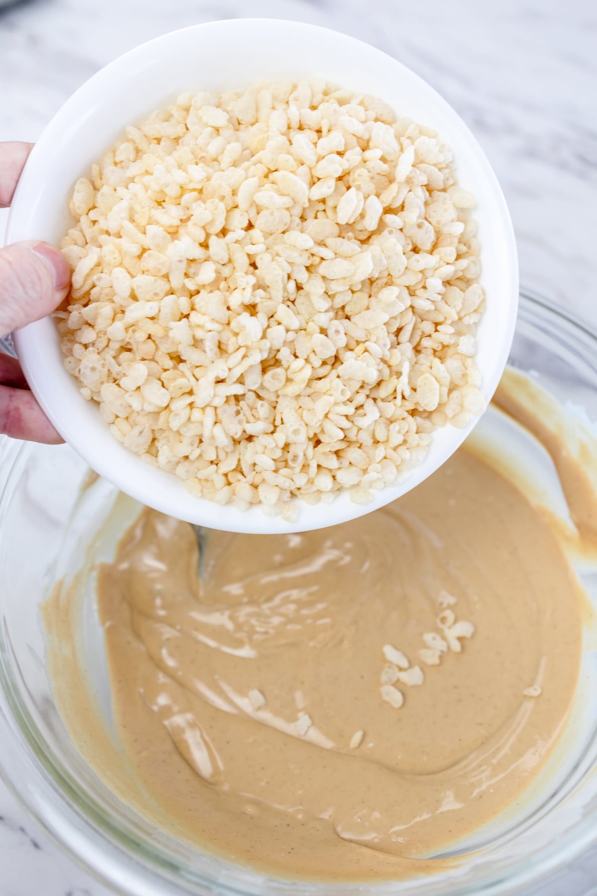 Top view of a white bowl of Rice Krispies about to be poured into a glass mixing bowl with peanut butter and almond bark mixture in it.