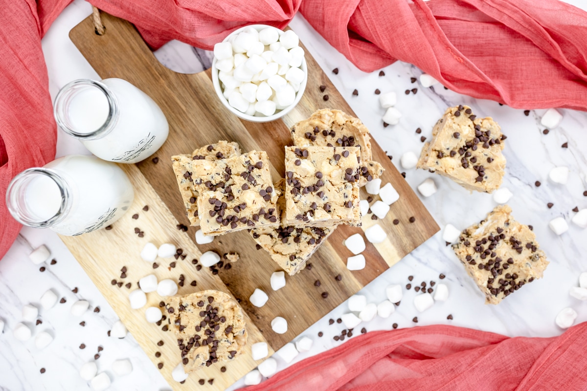 Top view of stacked Avalanche Bars on a wooden chopping board surrounded by bottles of milk, and additional marshmalloes and chocolate chips, boardered by red tea towels.
