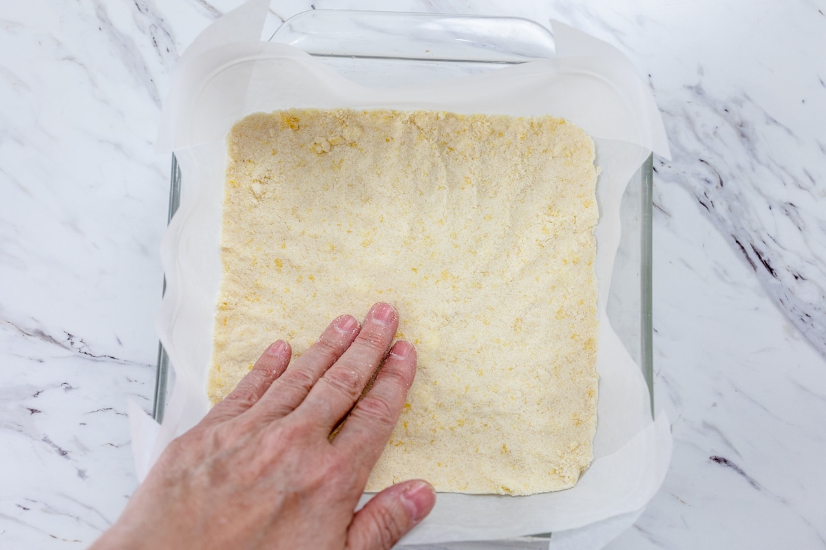 Top view of a square glass dish lined with parchment paper with crumble mixture in the bottom being pressed down by a hand.