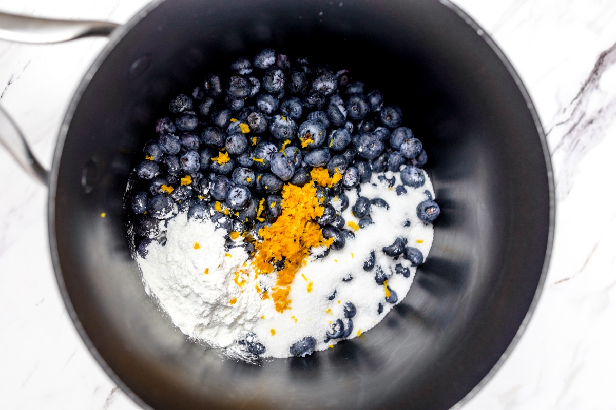 Top view of a black saucepan with blueberries, orange zest, and sugar in it.