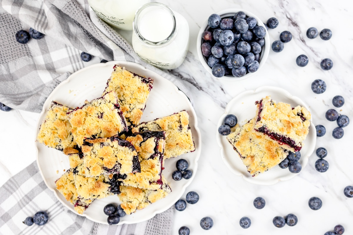 Top view of blueberry pie bars on a large plate, next to blueberry pie bars on a small plate, with a glass bottle of milk and a small bowl of blueberries next to it, surrounded by blueberries.
