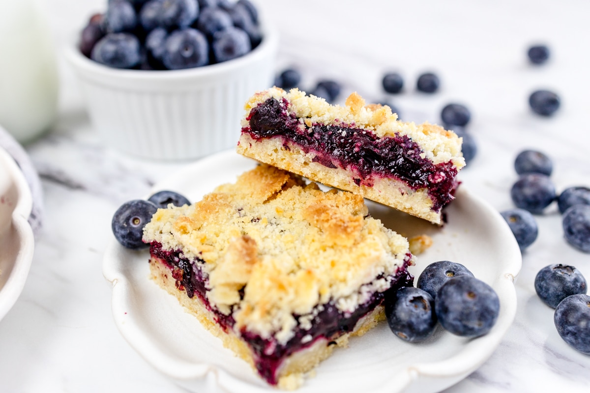 Close up of two blueberry pie cookie bars on a white plate, surrounded by blueberries.