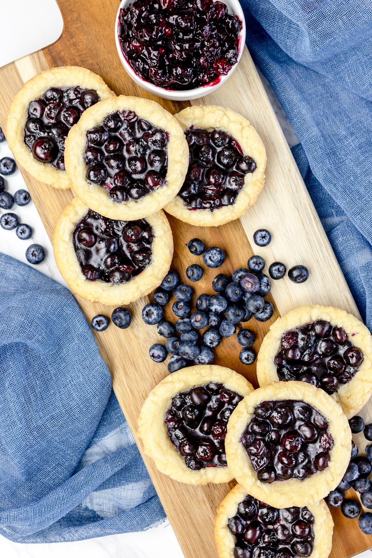 Top view of blueberry pie cookies on a serving board on a table with blue teatowels and blueberries next to them. 