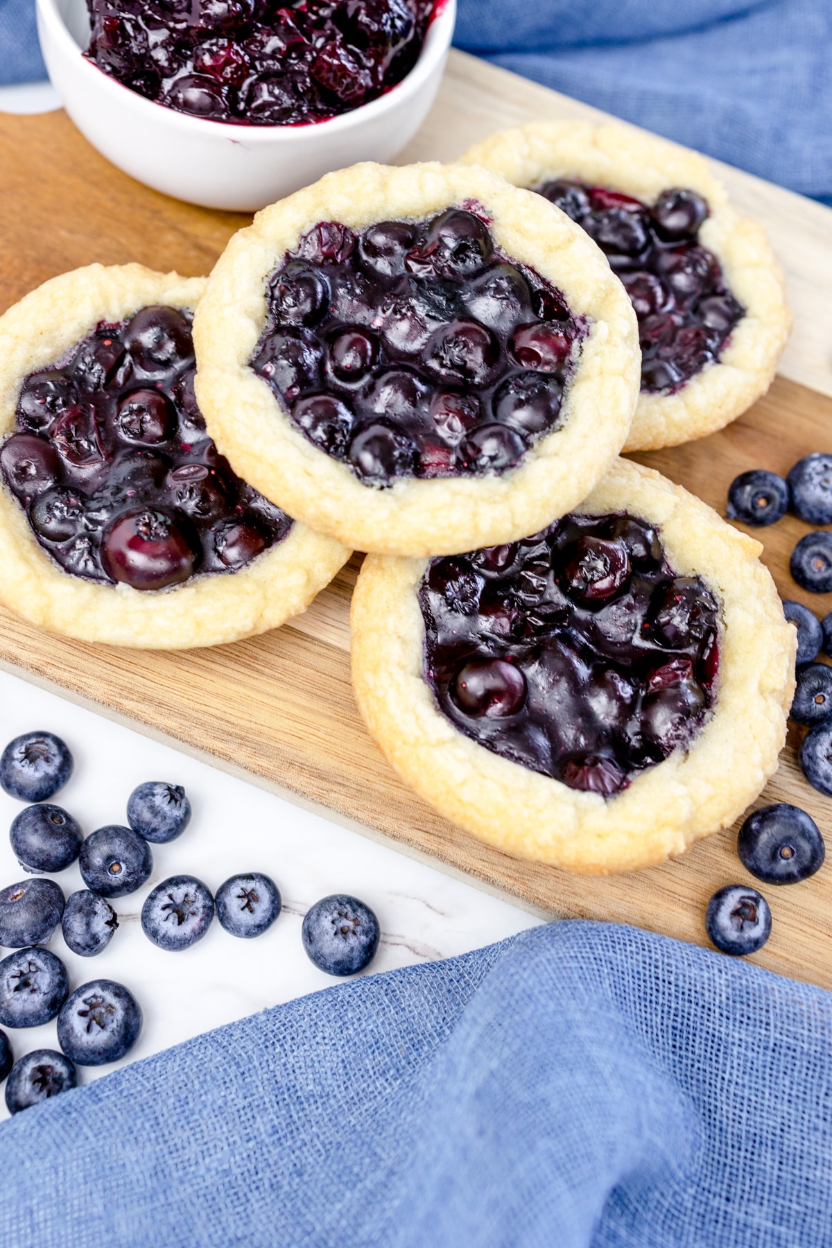 Close up of blueberry pie cookies on a serving board on a table with blue teatowels and blueberries next to them. 