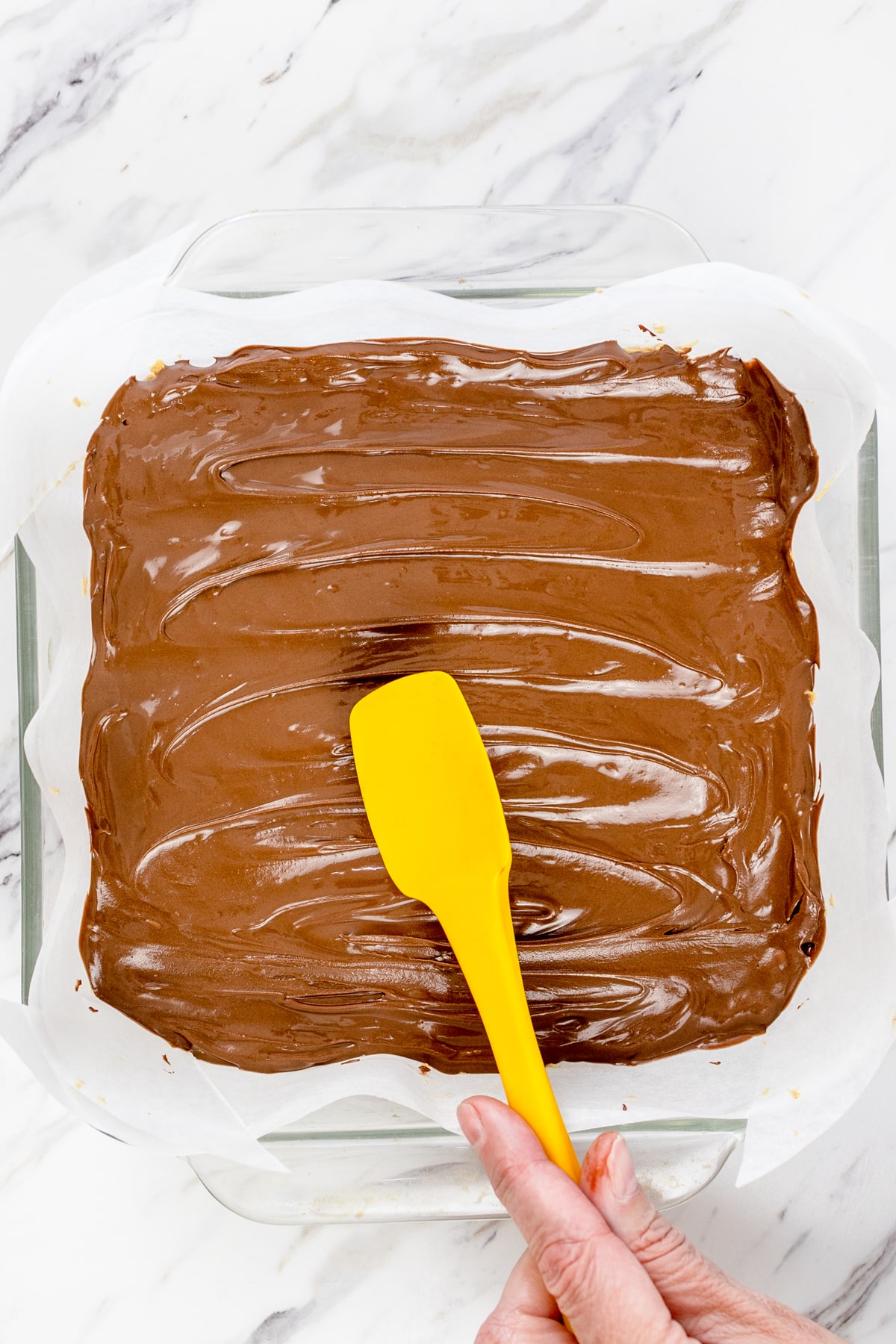 Top view of a square baking dish lined with parchment paper with a peanut butter mixture in the bottom with a melted chocolate mixture on top being spread out by a spatula.