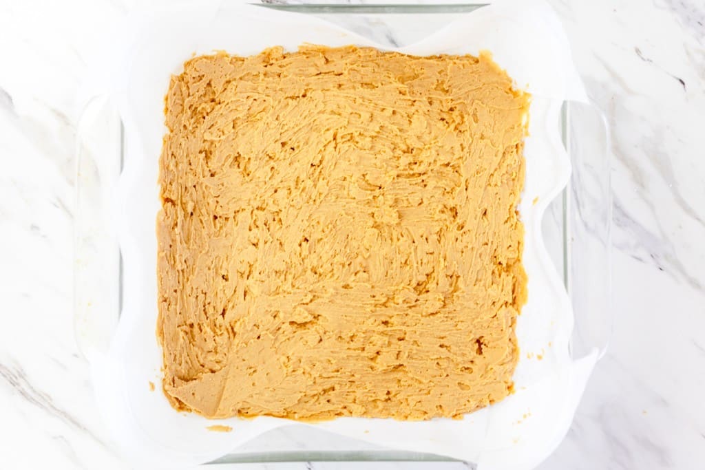 Top view of a square baking dish lined with parchment paper with a peanut butter mixture in the bottom of a baking dish. 