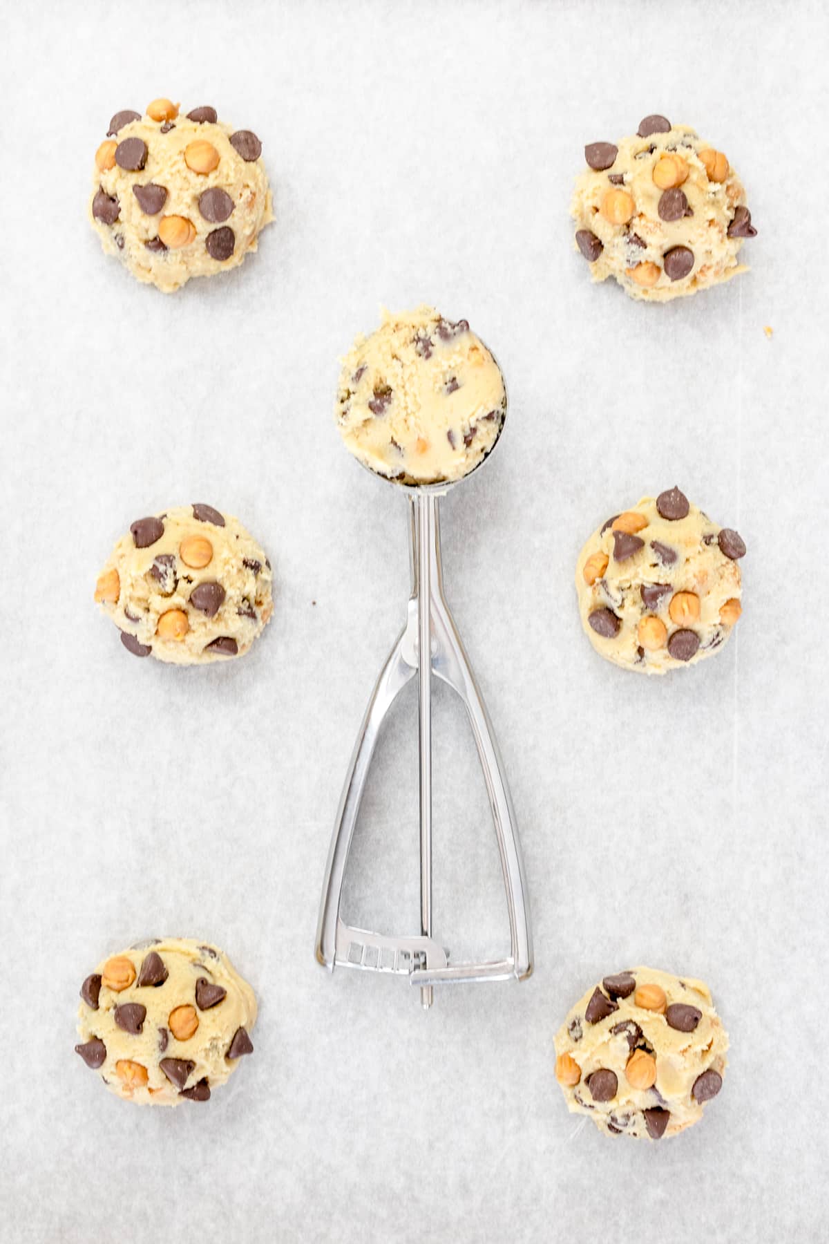 Top view of a cookie dough scoop filled with chocolate chip and caramel cookie dough surrounded by other cookie dough balls on a baking tray lined with parchment paper.
