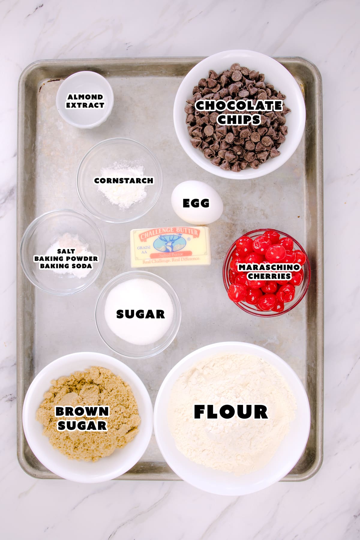 Top view of the ingredients needed to make chocolate chip cherry cookies in small bowls on a baking tray. 