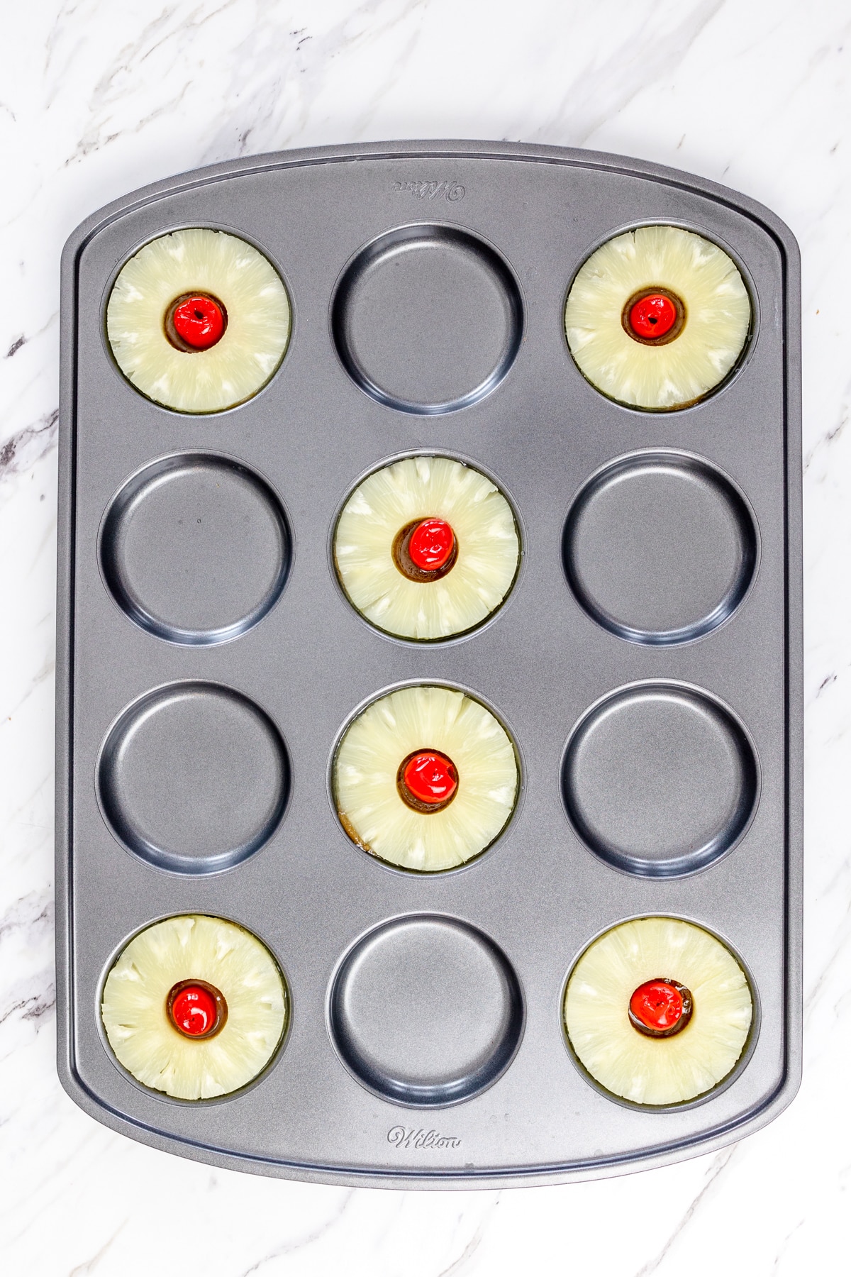Top view of a muffin top baking tray with pineapple rings in the cups and maraschimo cherries in the middle of them.