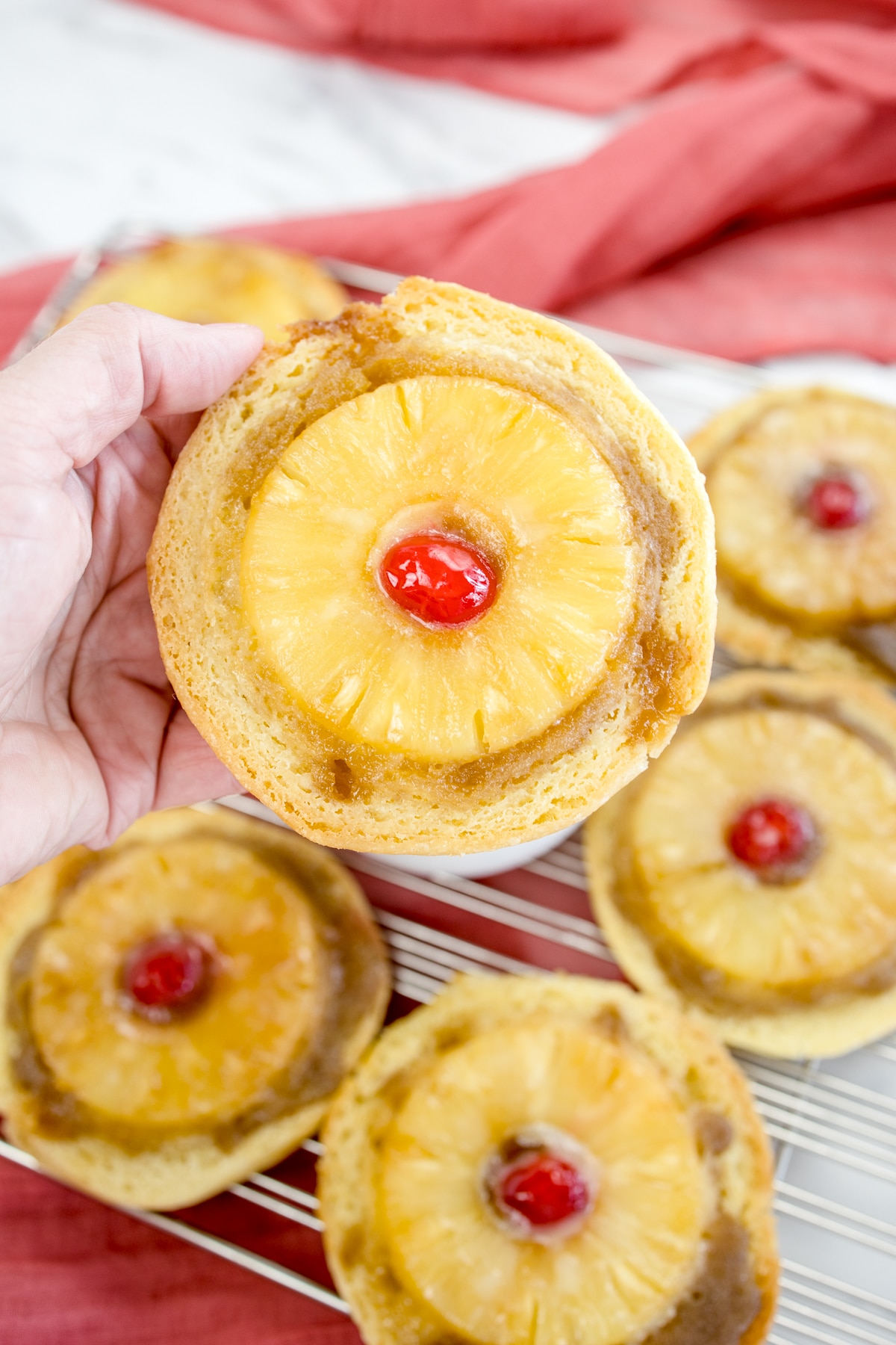 Pineapple Upside Down Cookies with a maraschino cherry in the middle on a wire rack, with one cookie being held in mid-air by a hand.