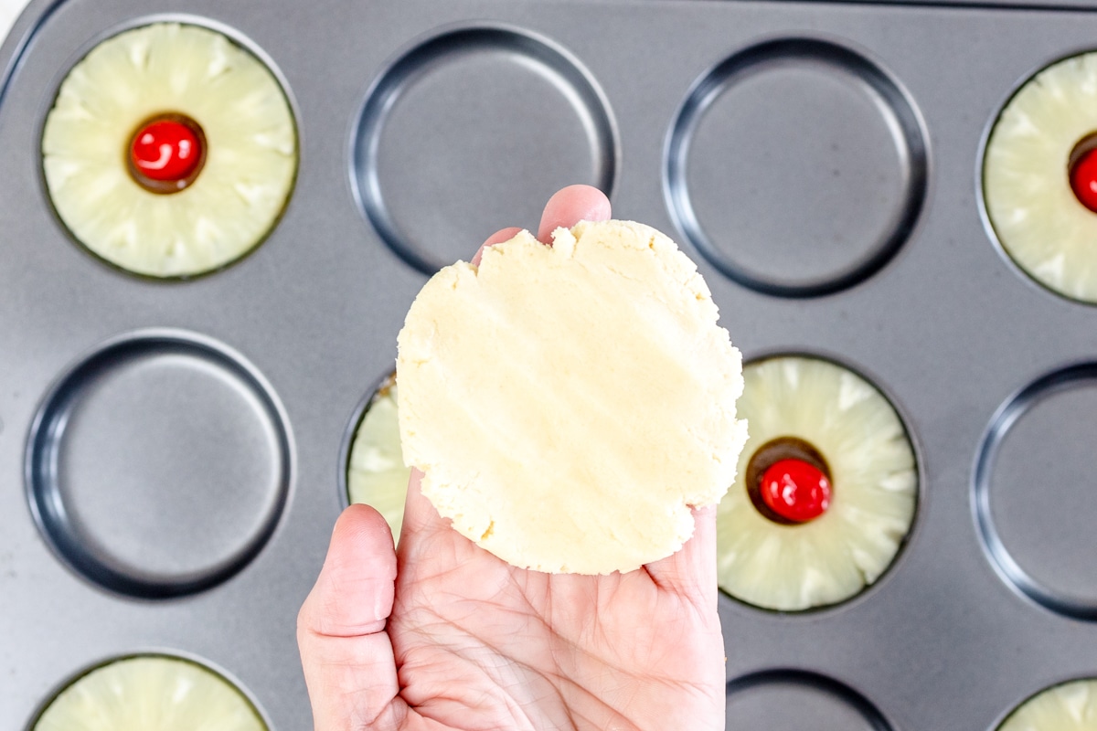 Top view of a hand holding a flattened cookie dough circle in mid-aiir above a muffin top baking tray with pineapple rings and cherries in it.