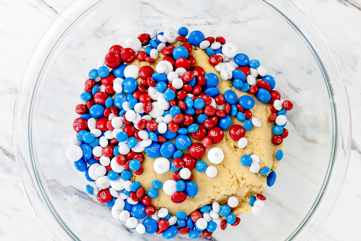 Top view of a glass mixing bowl with cookie dough and red, white, and blue M&Ms on top.
