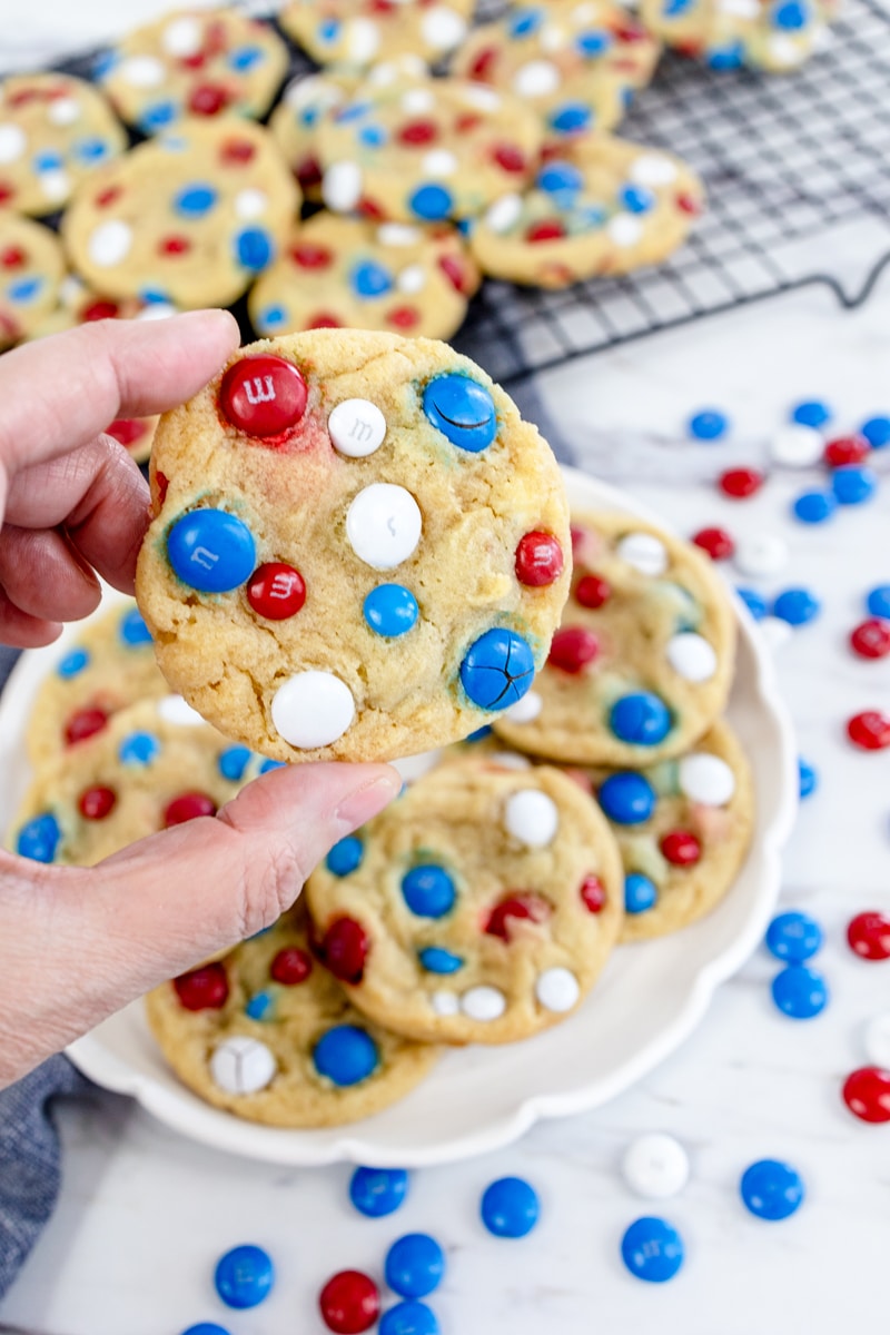 Close up of 4th of July Cookies on a white plate surrounded by red, white, and blue M&Ms, with one cookie being lifted off the plate.