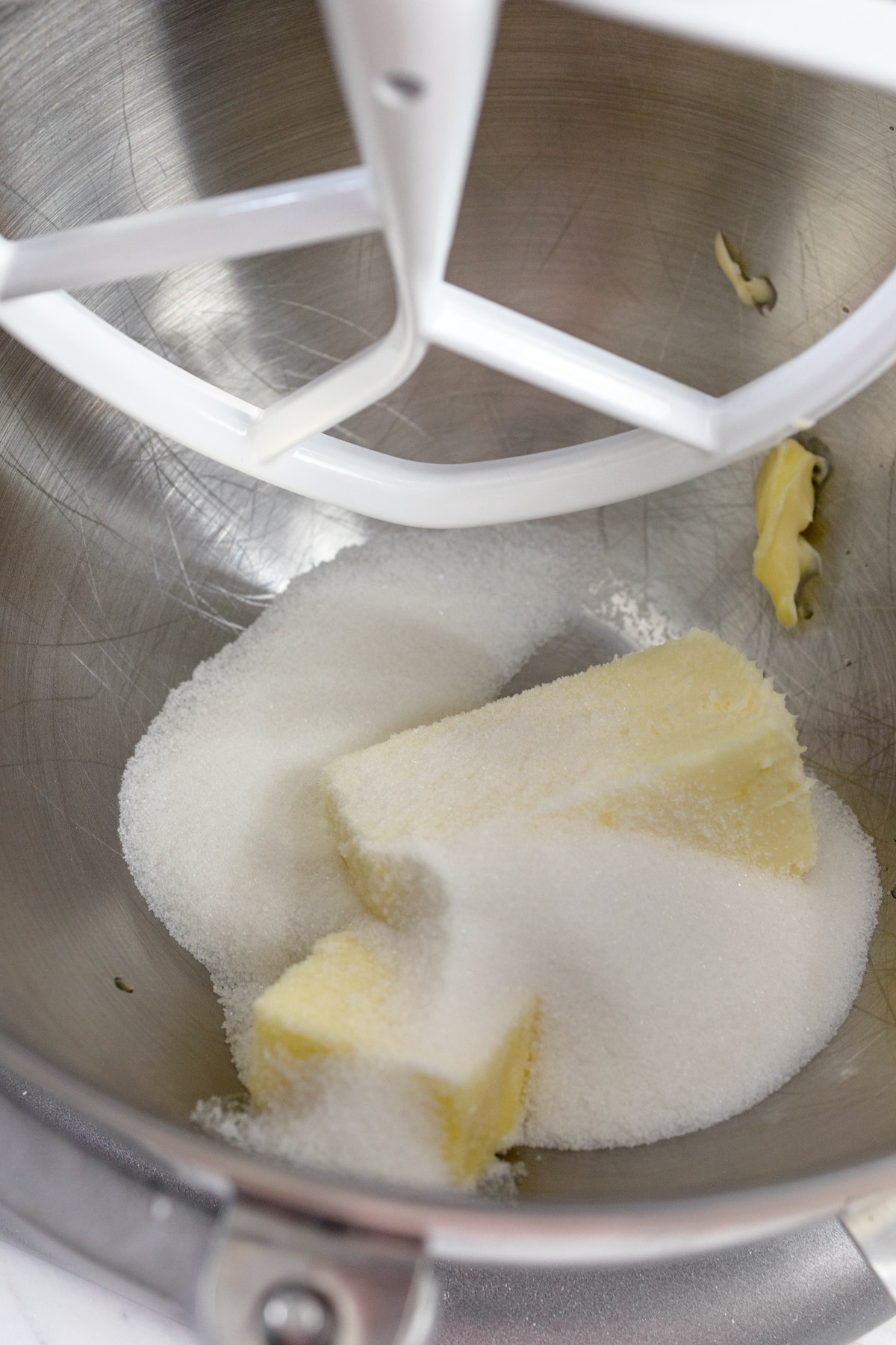 Close up of the bowl of a stand mixer with blocks of butter and white sugar in it with a paddle attachment.