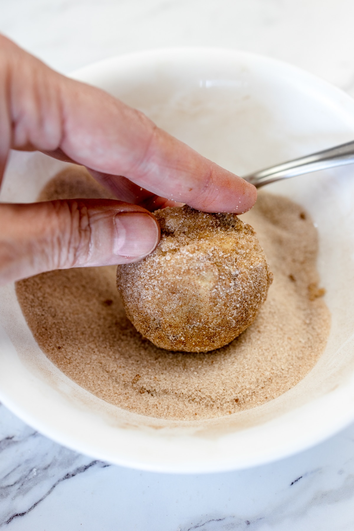 Close up of a cookie dough ball being rolled into a small bowl with cinnamon sugar in it.