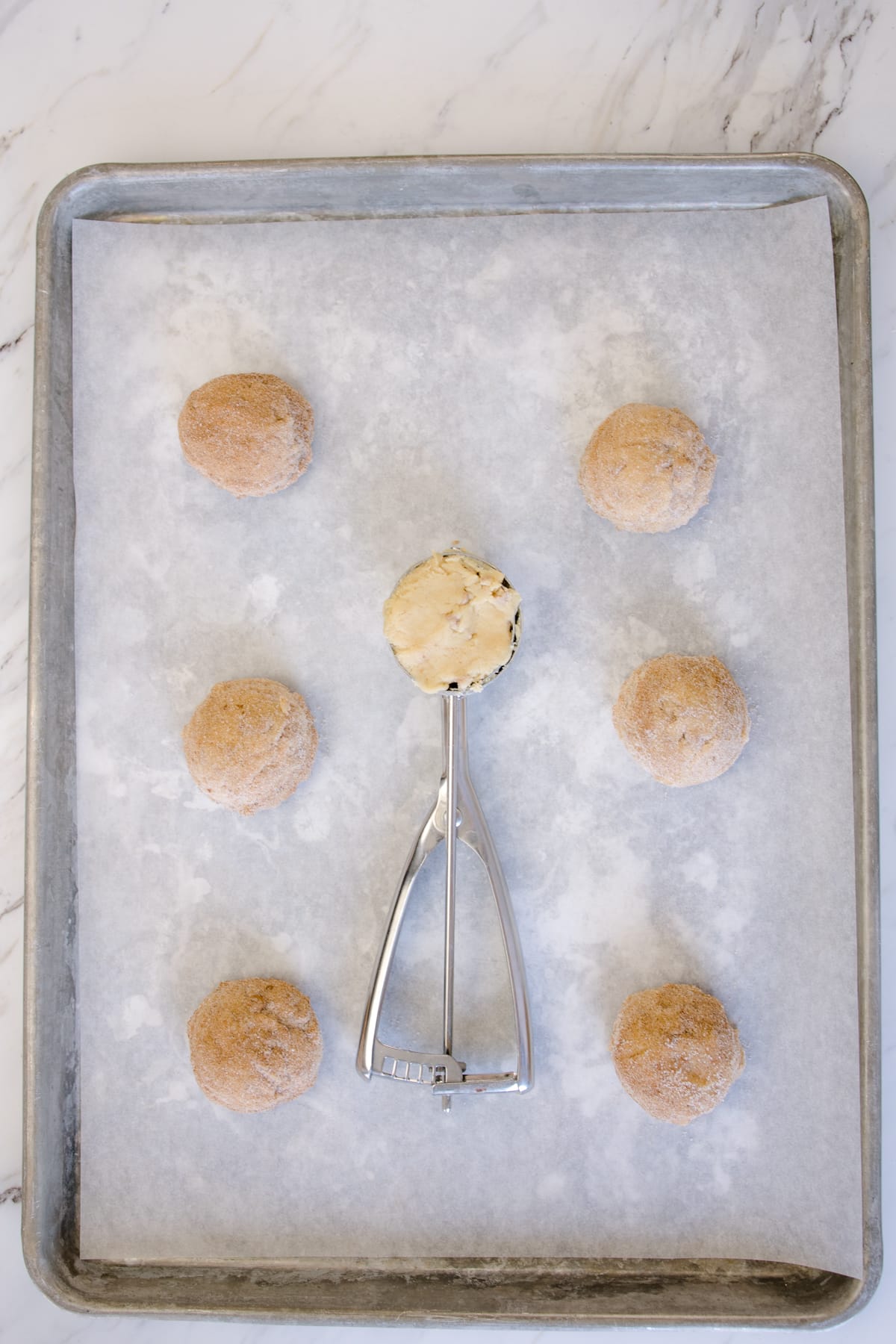 Top view of baking tray lined with parchment paper with Cinnamon Chip Cookie dough balls on it, the middle one still inside the cookie scoop.