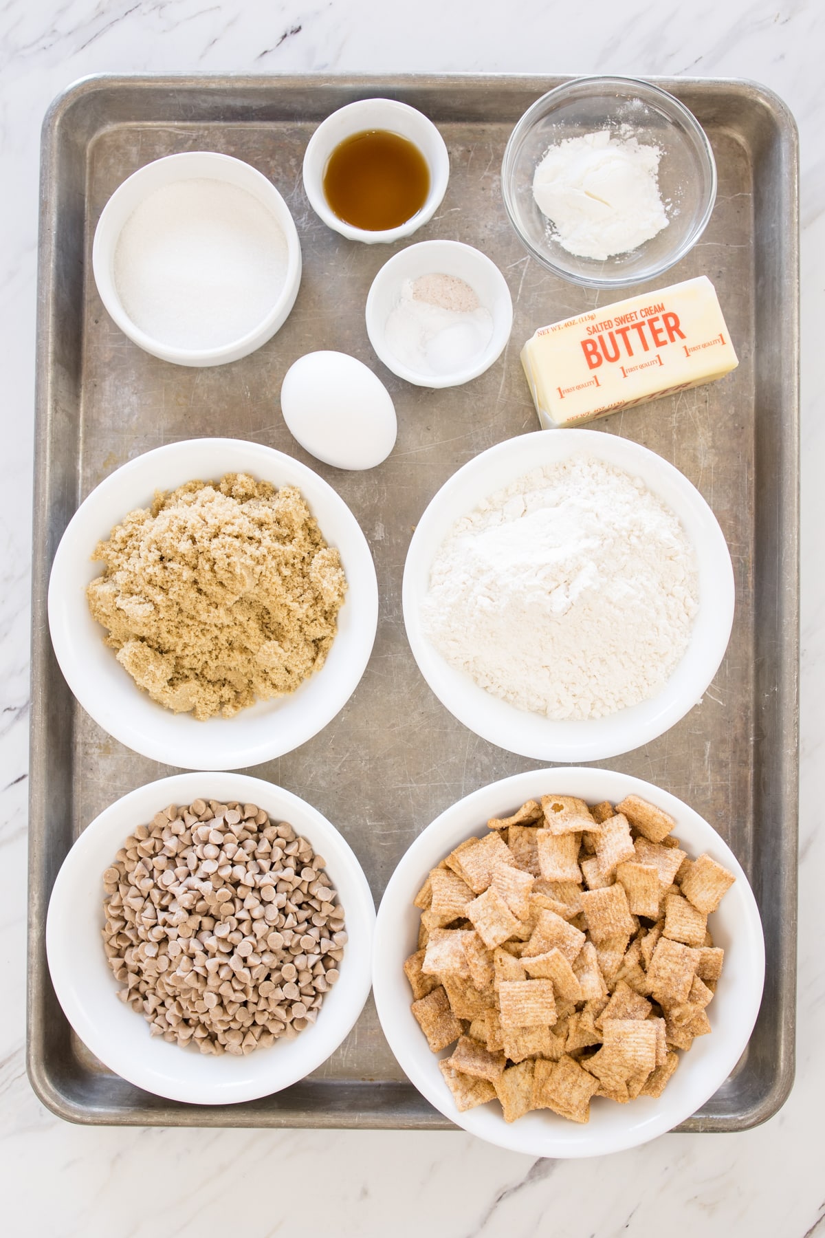 Top view of ingredients needed to make cinnamon toast crunch cookies in small bowls on a baking tray.