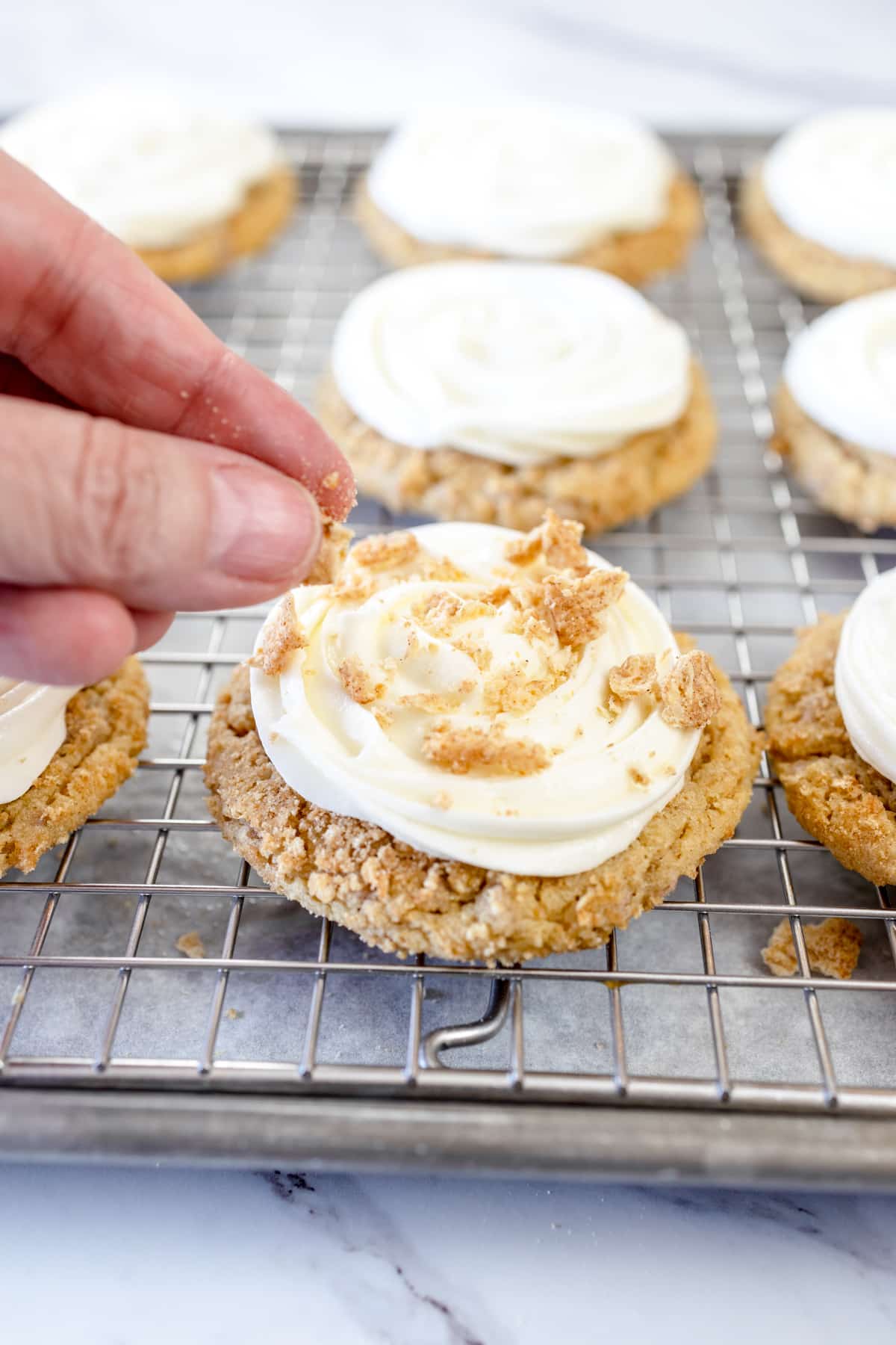 Close up of frosted cinnamon toast crunch cookies on a wire rack being topped with a cinnamon toast crunch topping.