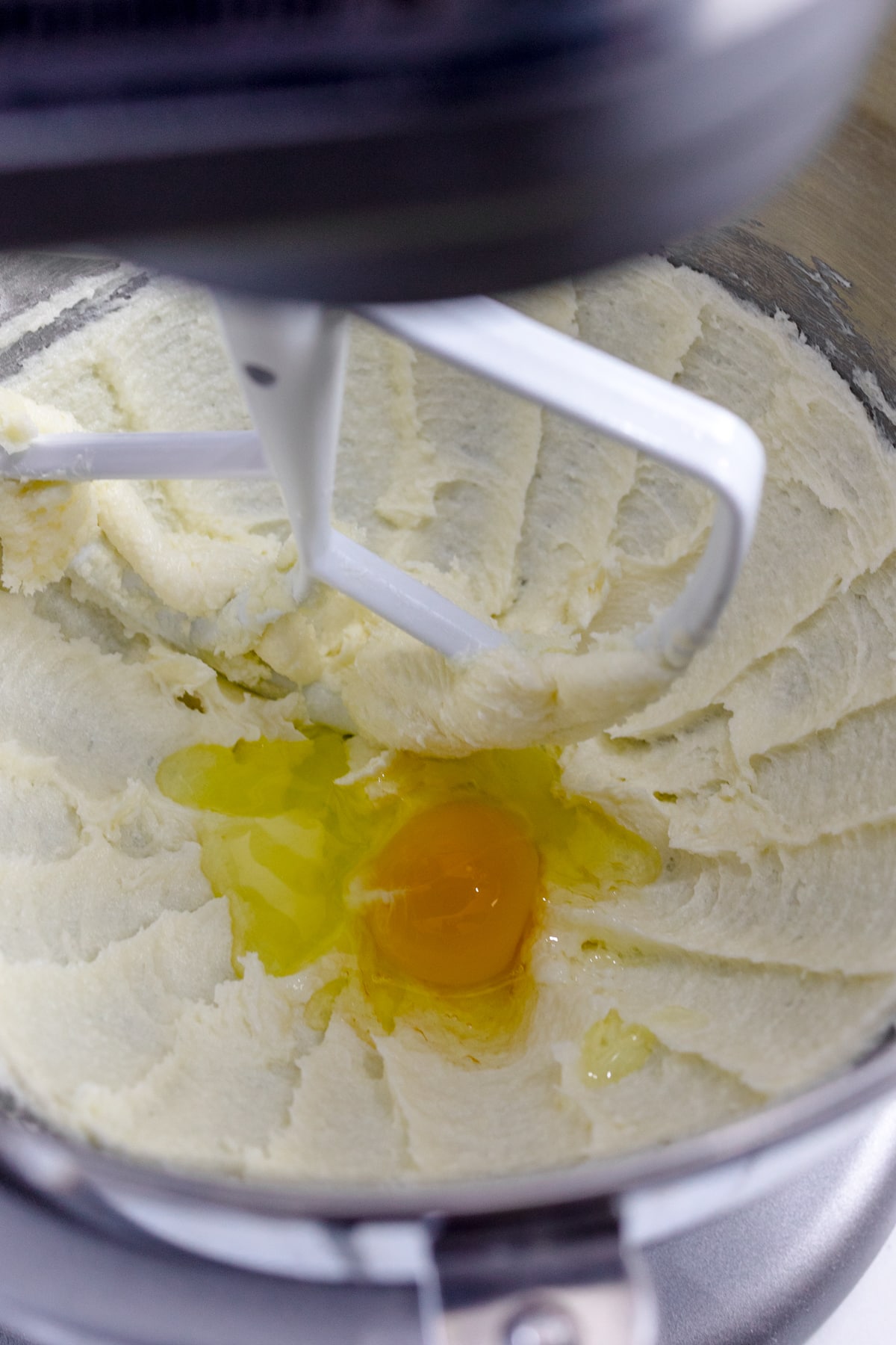 Close up of a bowl of a stand mixer with a paddle attachment with a creamed butter and sugar mixture in the bottom and an egg on top.