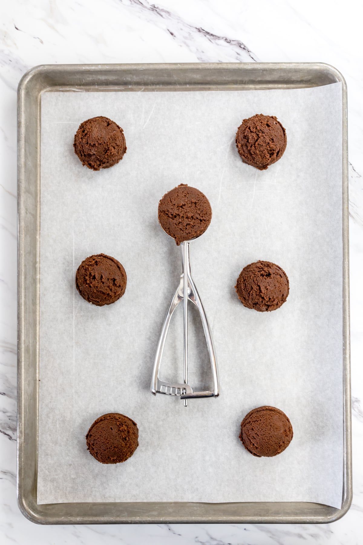 Top view of a baking tray lined with parchment paper with chocolate cookie dough balls on it, and a cookie scoop lying face up with a chocolate cookie scoop in it.