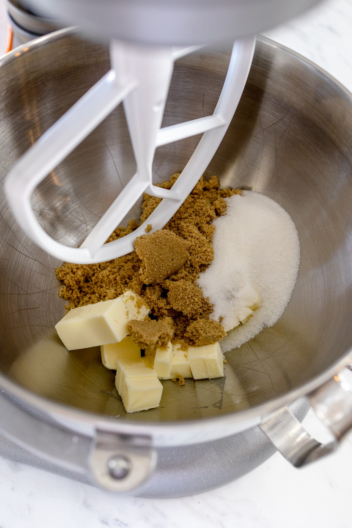 Close up of the bowl of a stand mixer with butter and sugars in it being mixed with a paddle attachment. 