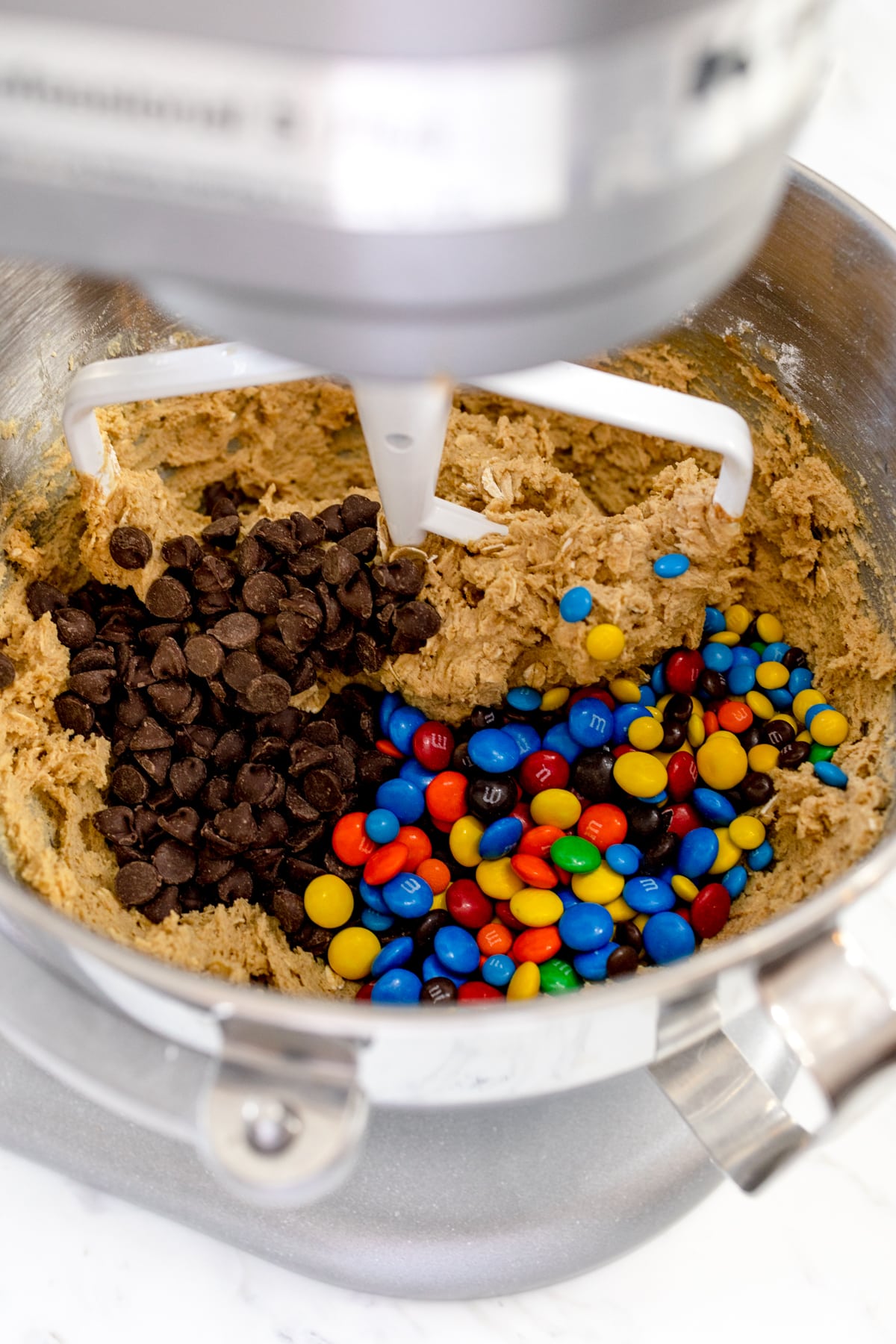Close up inside the bowl of a stand mixer with cookie dough in the bottom, and chocolate chips and M&Ms on top. 
