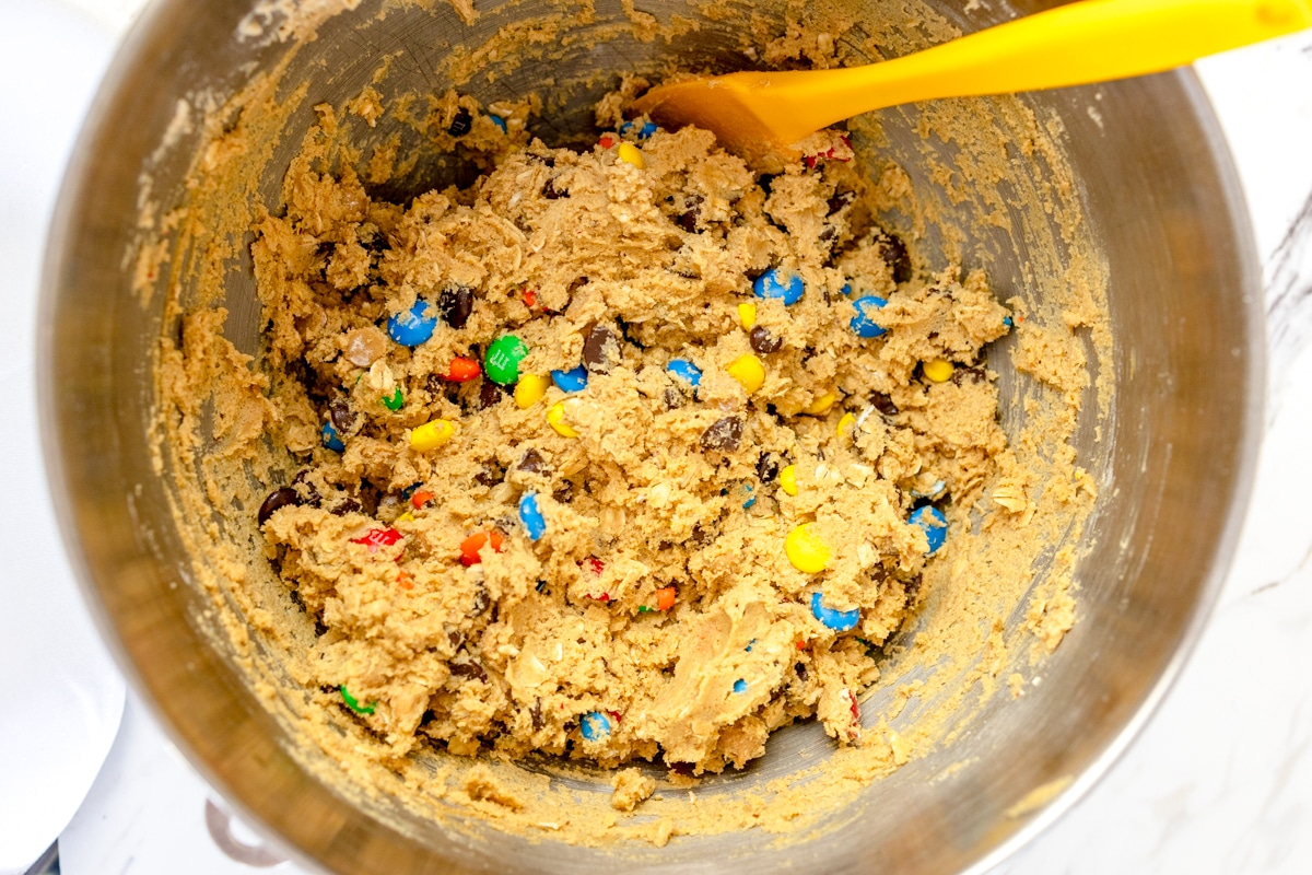 Top view of a metal mixing bowl with monster cookie bars cookie dough being mixed with a yellow spatula. 