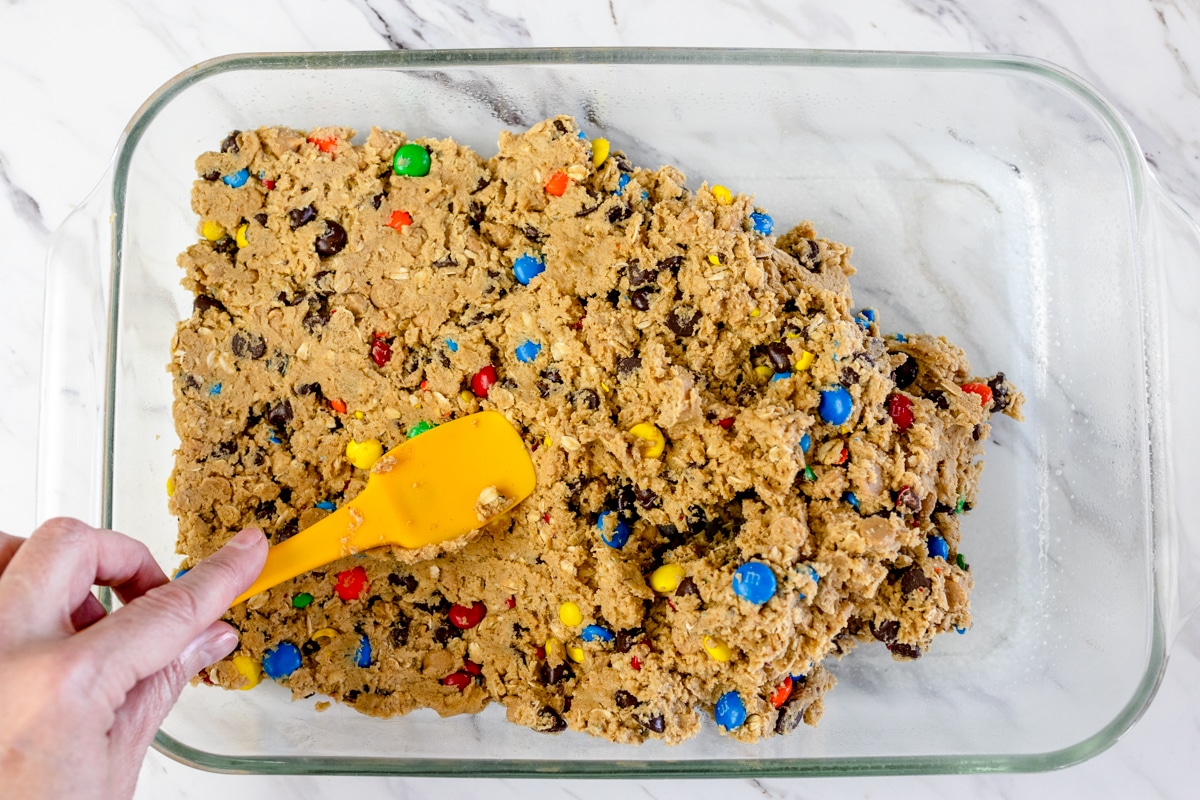 Top view of a glass baking dish with monster cookie bars cookie dough being pressed into the bottom of it with a yellow spatula.