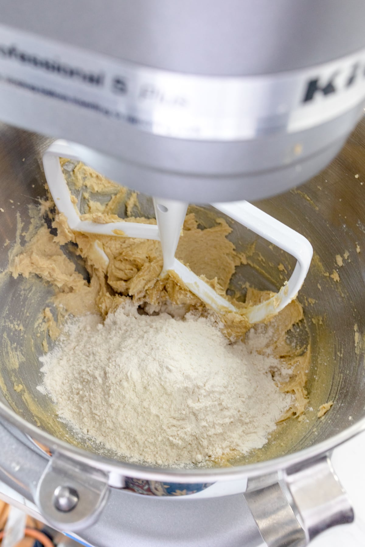 Close up of a stand mixer with a paddle attachment with a creamed mixture in it and a flour misture on top.