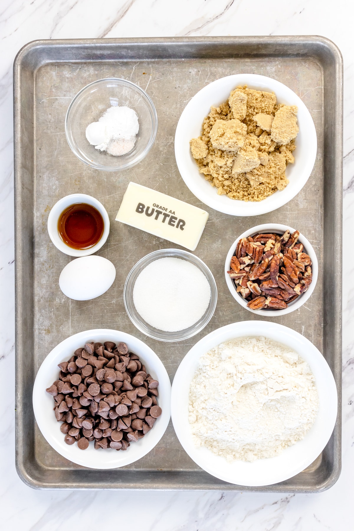 Top view of ingredients needed to make chocolate chip pecan cookies in small bowls on a baking tray.