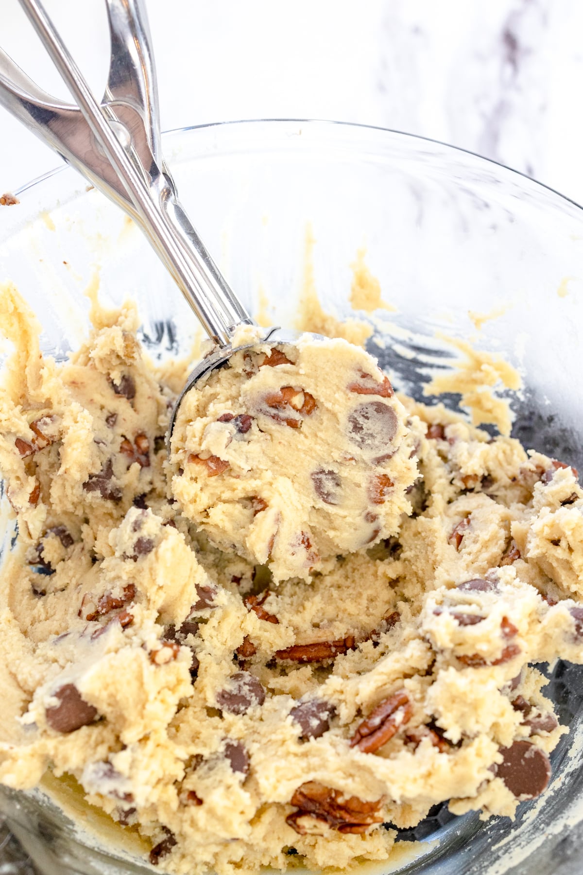 Top view of a glass bowl with chocolate chip pecan cookie dough being scooped out with a cookie scoop.