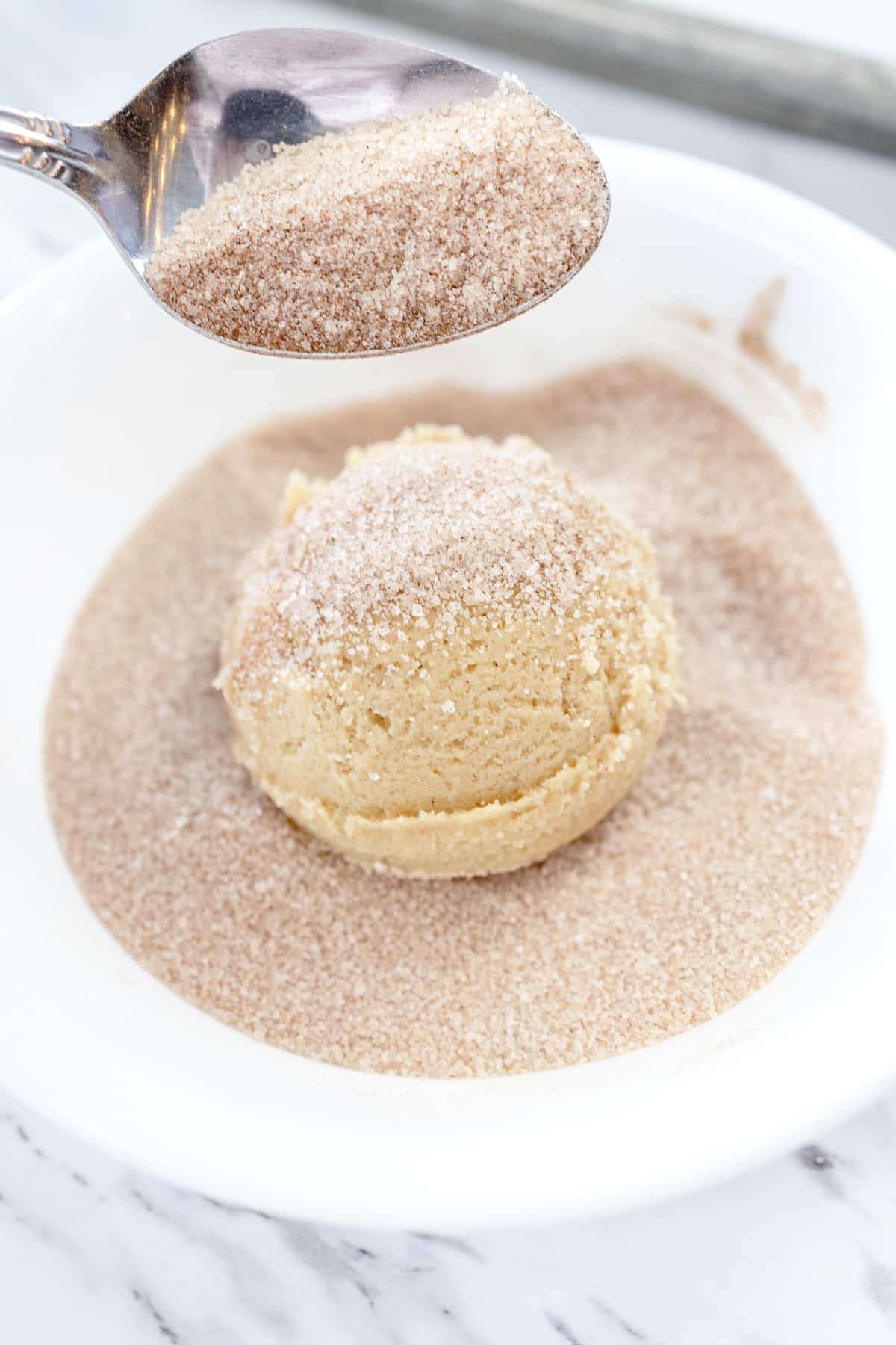 Close up of a ball of cookie dough being rolled in a bowl of cinnamon sugar.