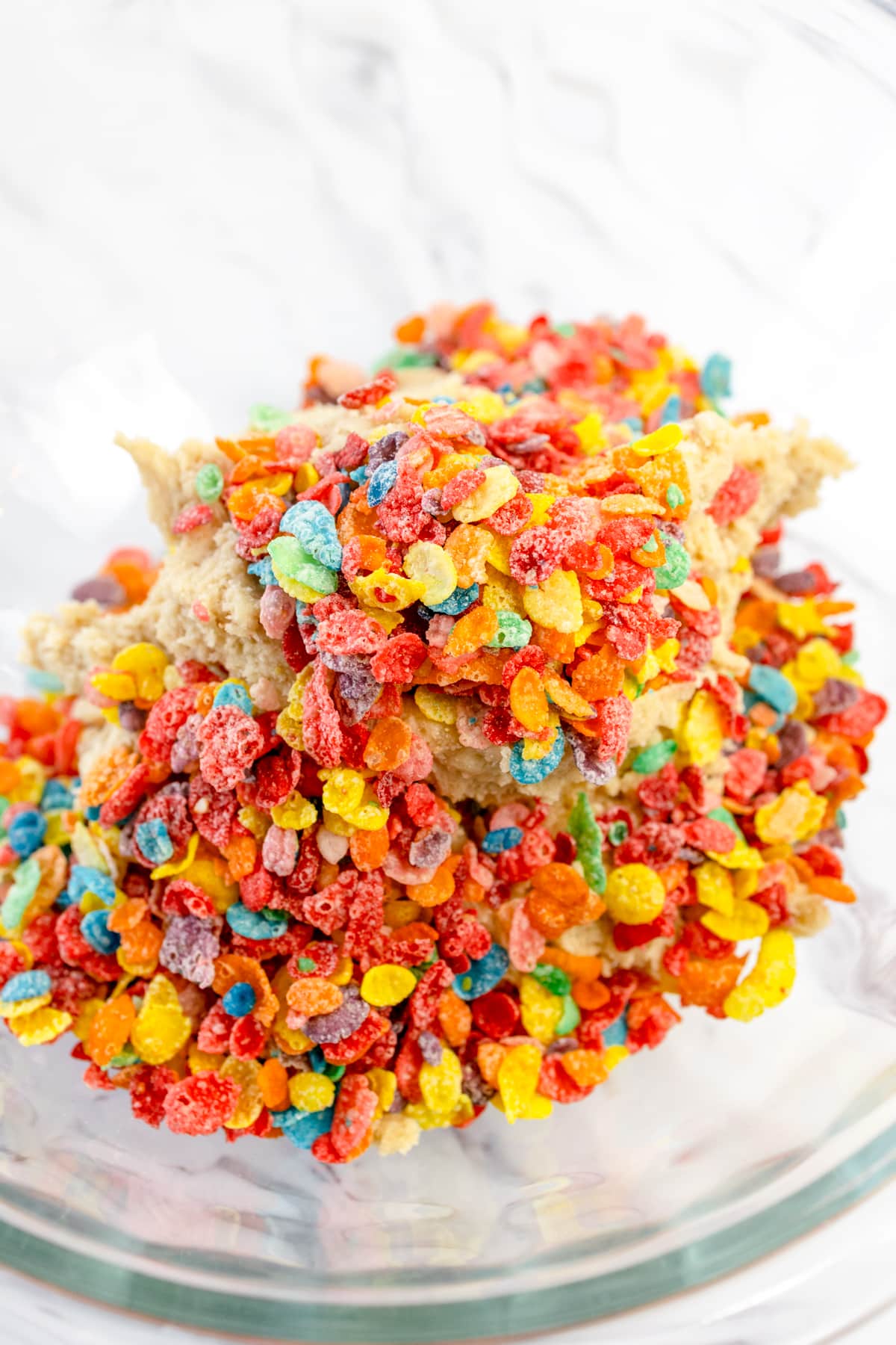 Top view of a glass mixing bowl with plain cookie dough in the bottom and fruity pebble cereal on top.